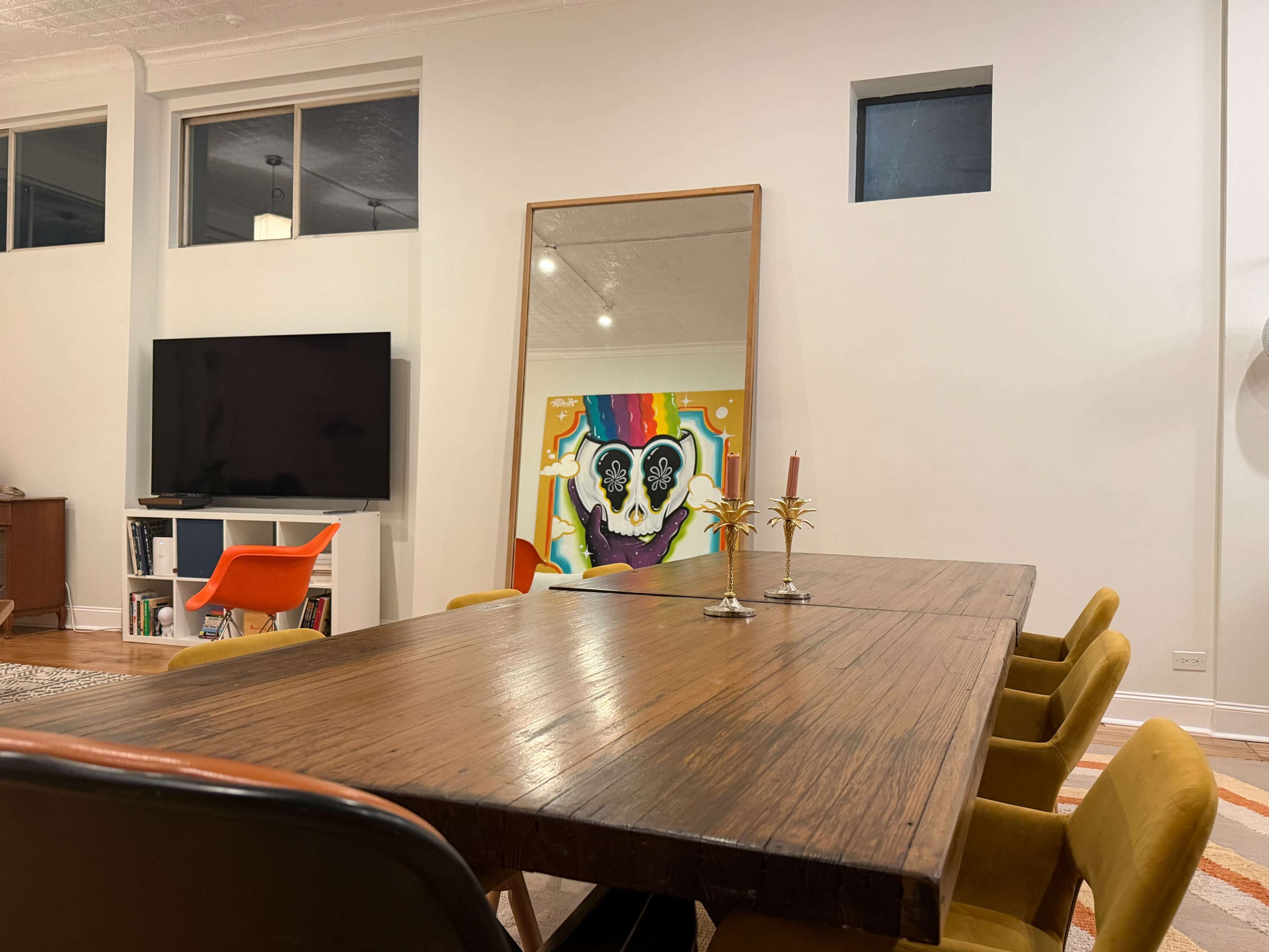 A large wooden dining table with yellow chairs is set in a well-lit room featuring a television and a colorful piece of artwork hanging on the wall.