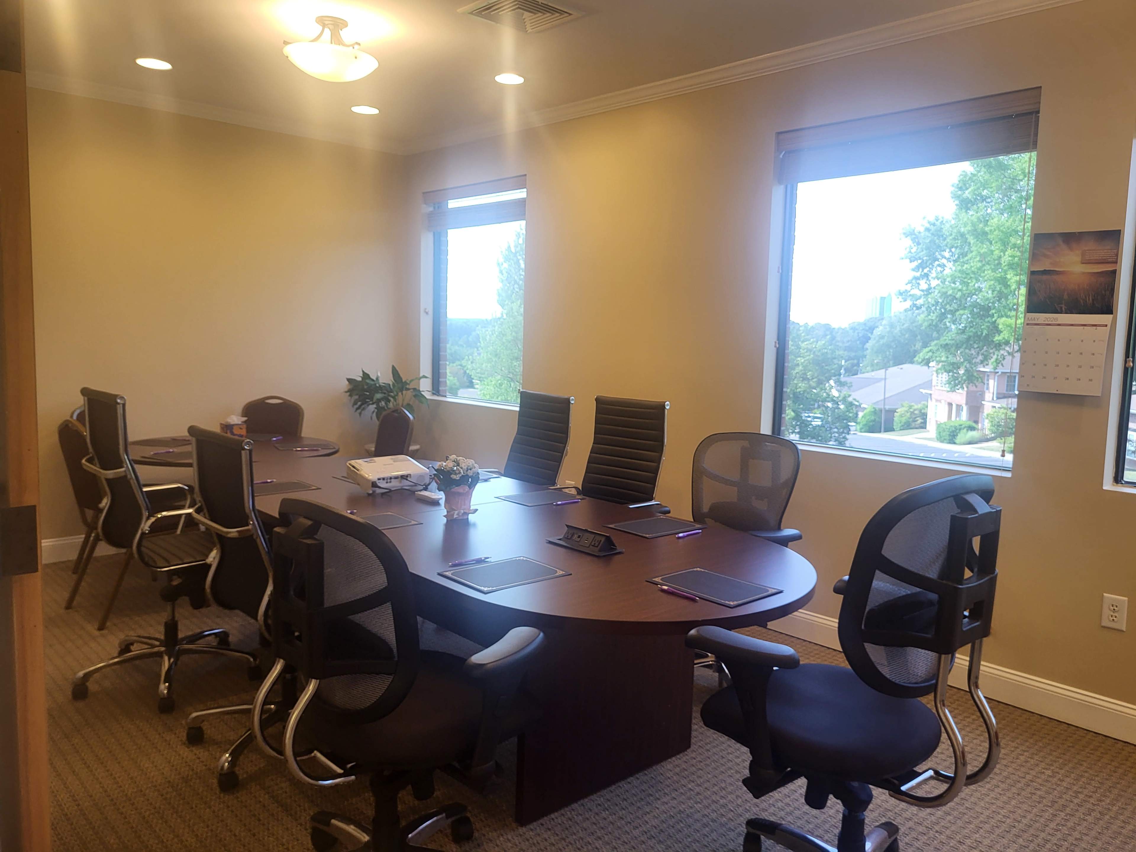 The image shows a modern conference room with a large round table surrounded by eight ergonomic chairs, two windows providing natural light, and a view of greenery outside.