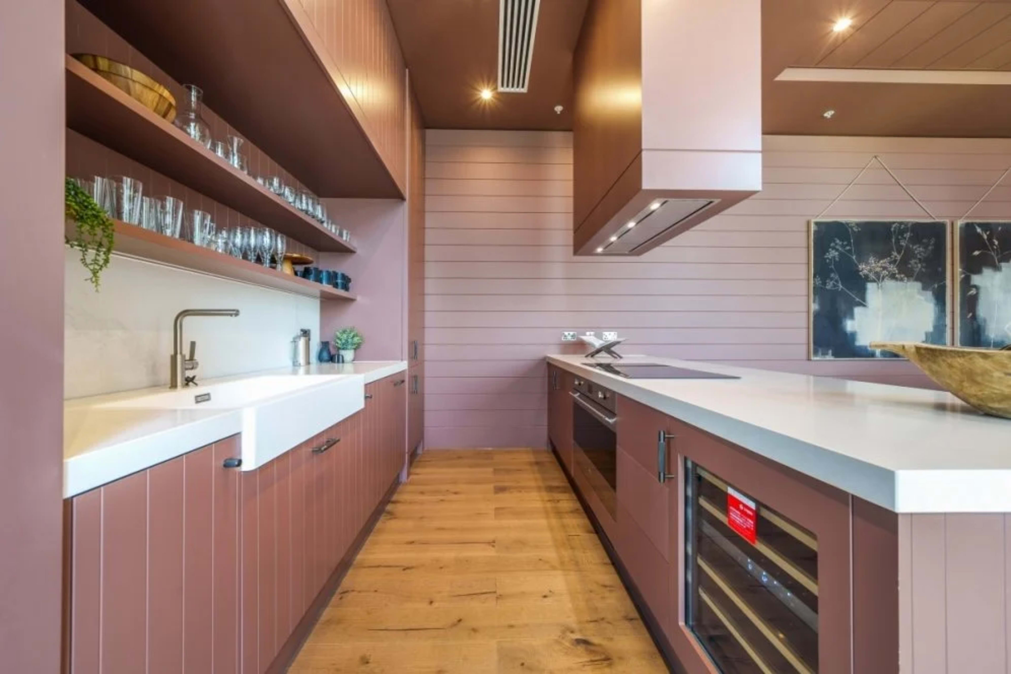 A modern kitchen with pink-hued cabinetry, open shelves displaying glassware, and a central island with a built-in stovetop.