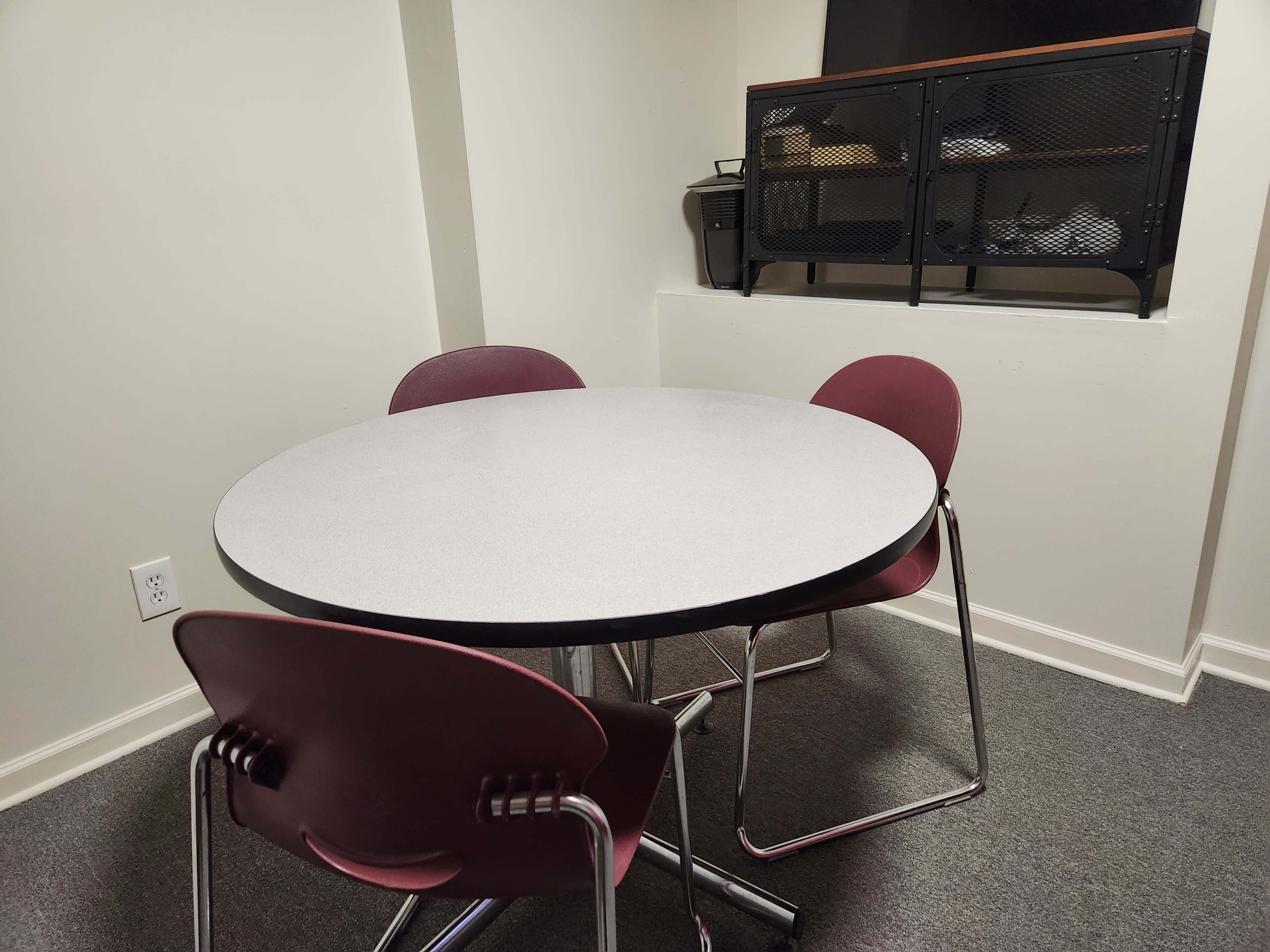 A round table with three burgundy chairs is positioned in a small room with gray carpeting, and a shelf is mounted on the wall behind it.