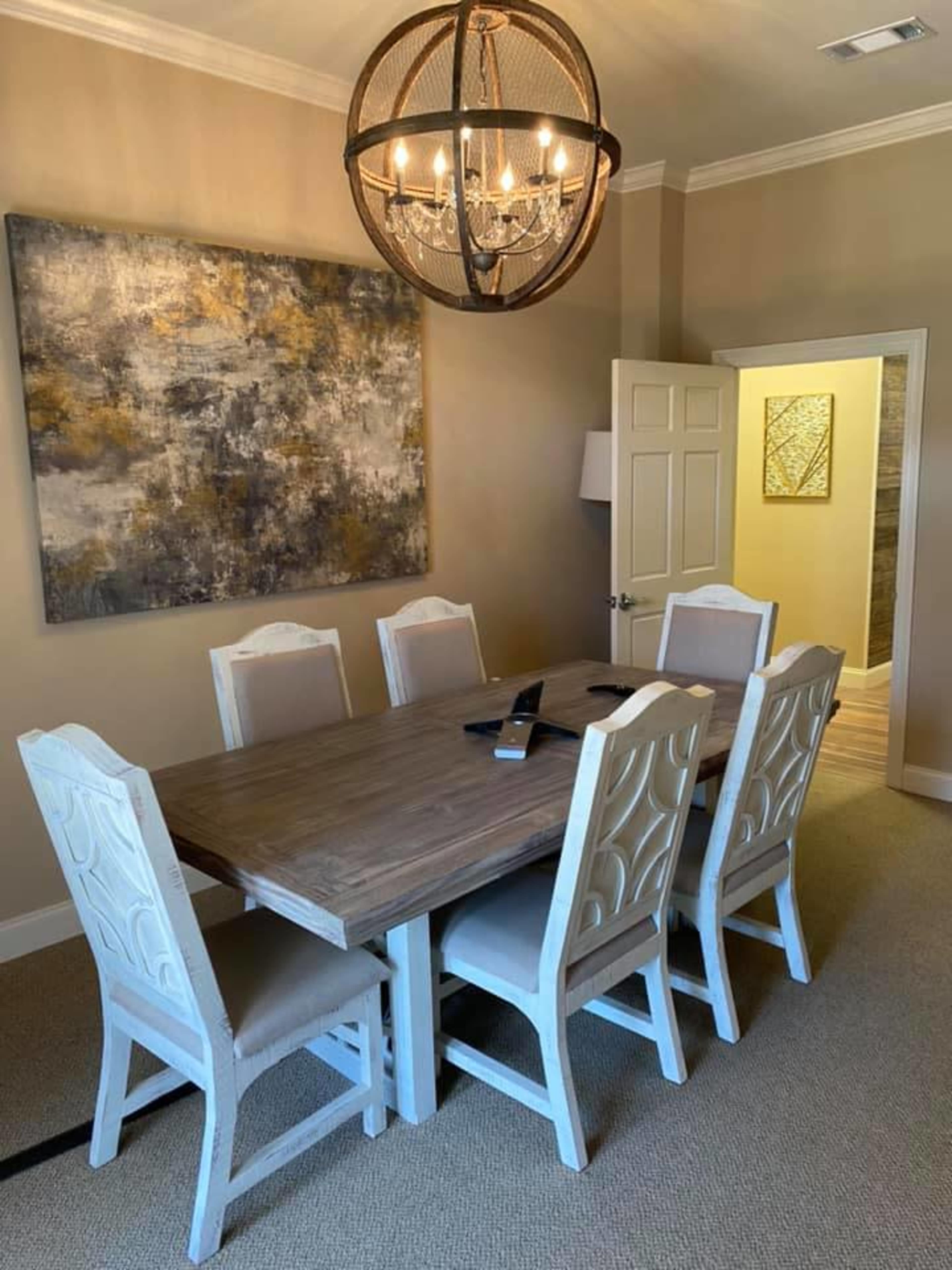 A dining room features a wooden table surrounded by six white chairs, with a large artwork on the wall and a pendant light above.