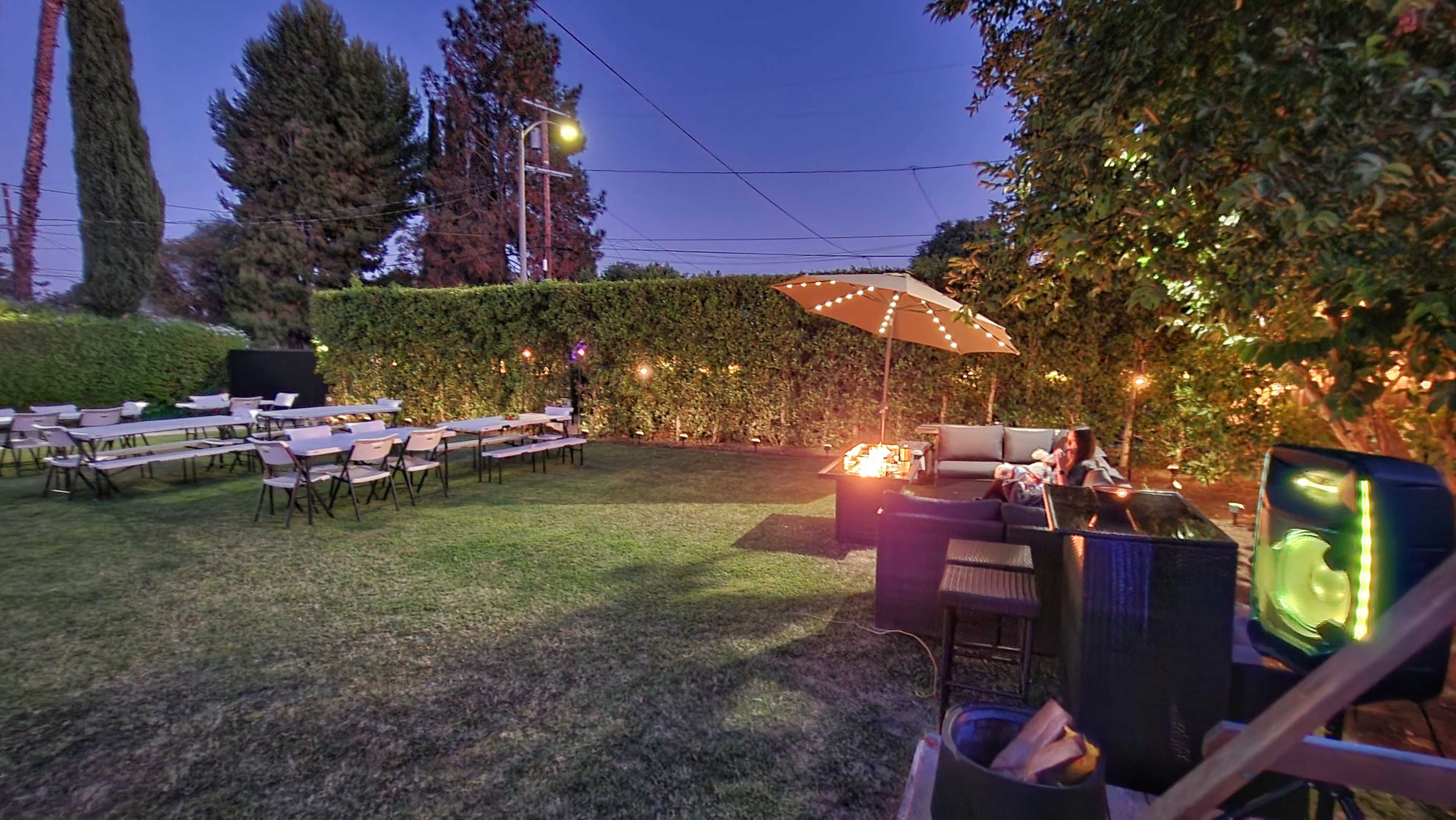 A backyard setting in the evening with neatly arranged tables, a fire pit, and an umbrella over a seating area surrounded by hedges and string lights.