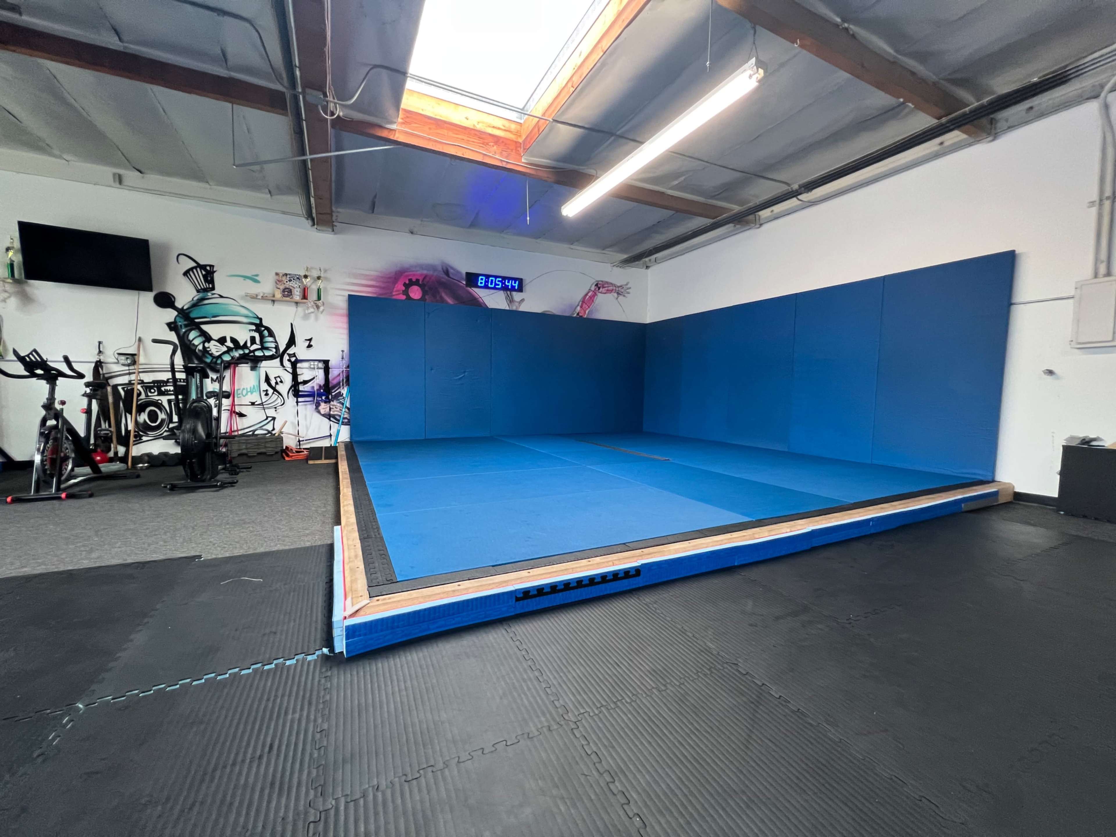 A gym space featuring a large blue mat area surrounded by black flooring and exercise equipment in the background.