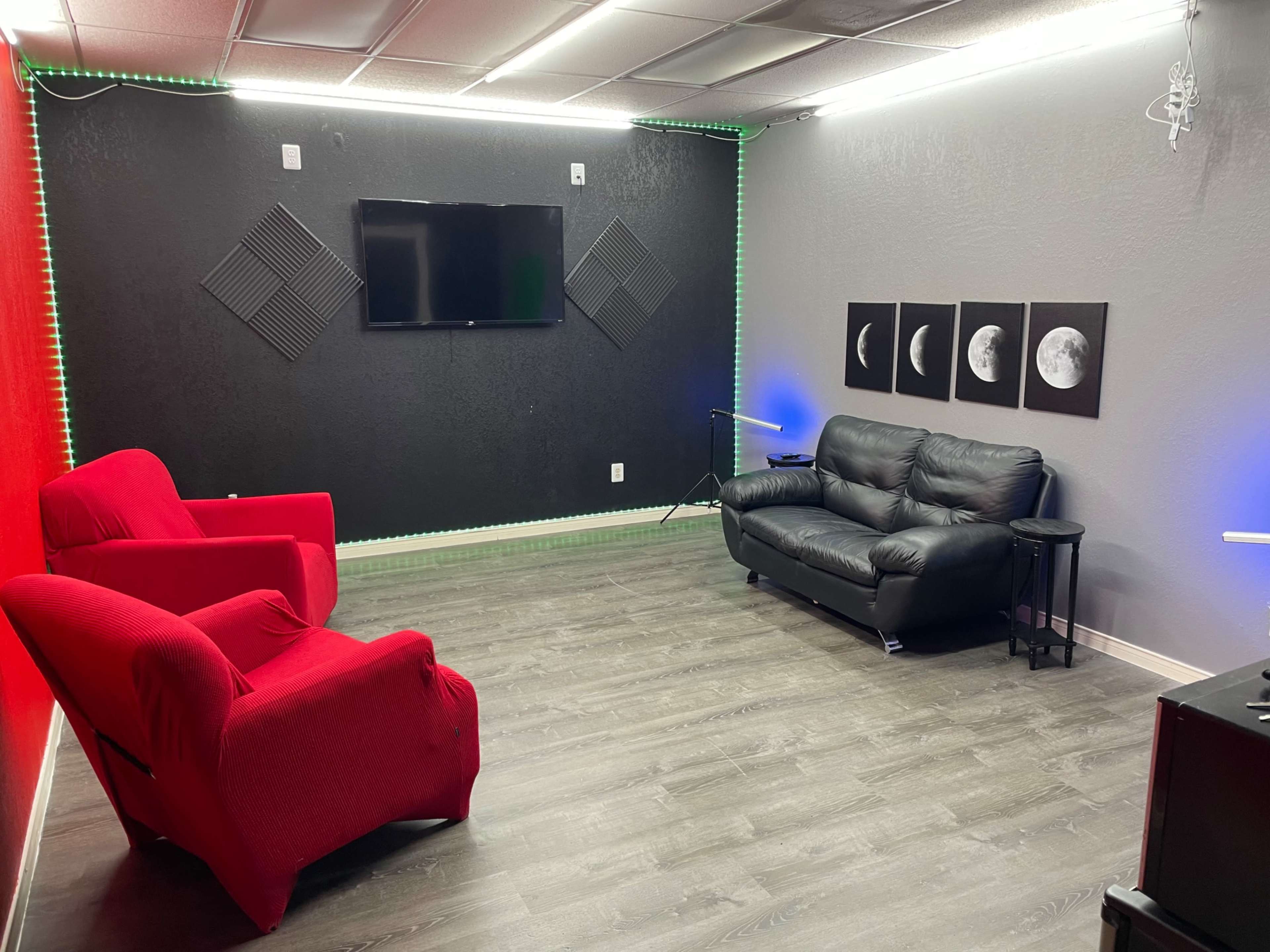 The room features a television mounted on the wall, a black sofa, two red chairs, and three framed lunar images.