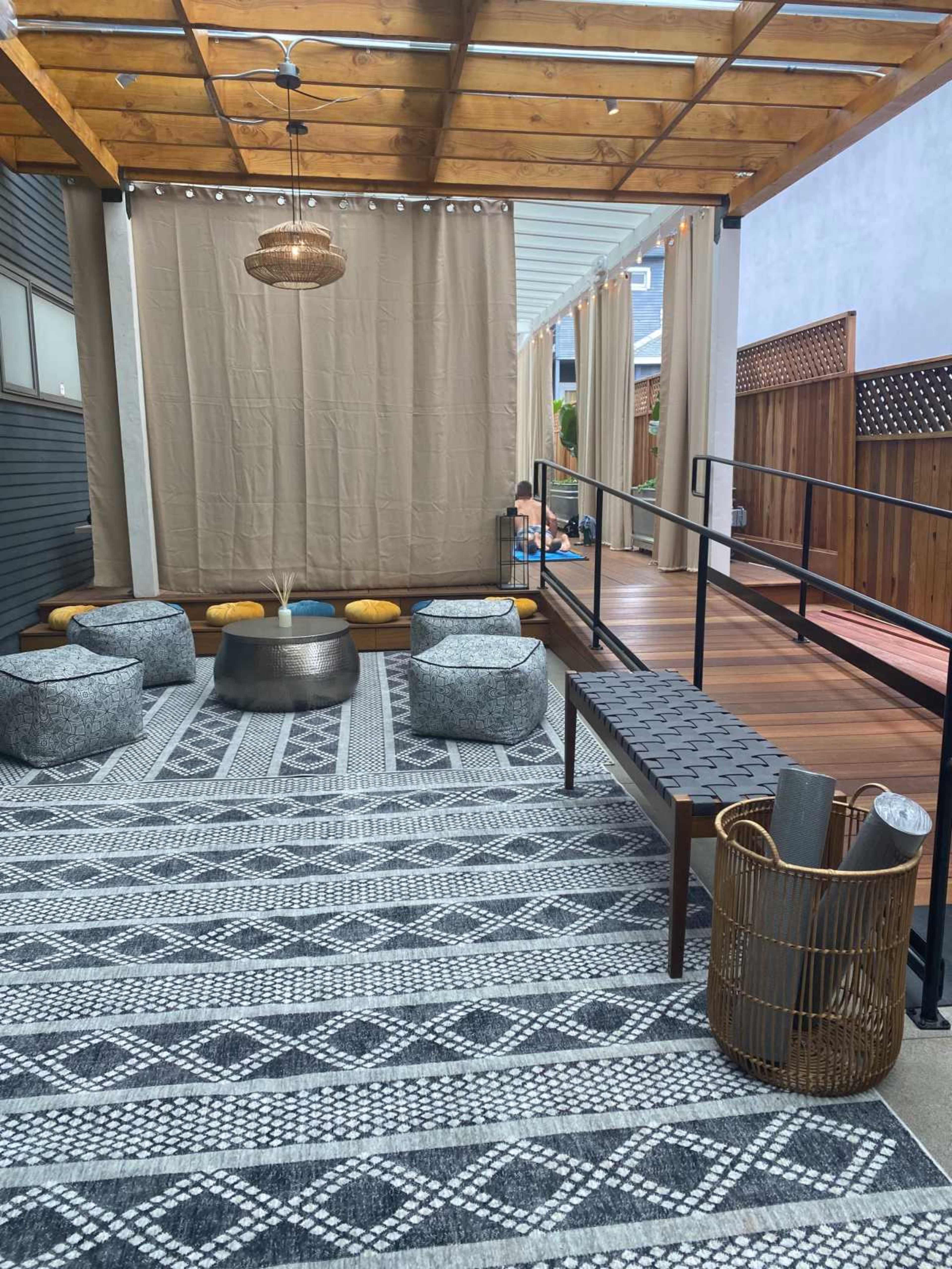 The image shows a modern outdoor seating area with patterned rugs, cushioned stools, a wooden bench, and a shaded walkway.