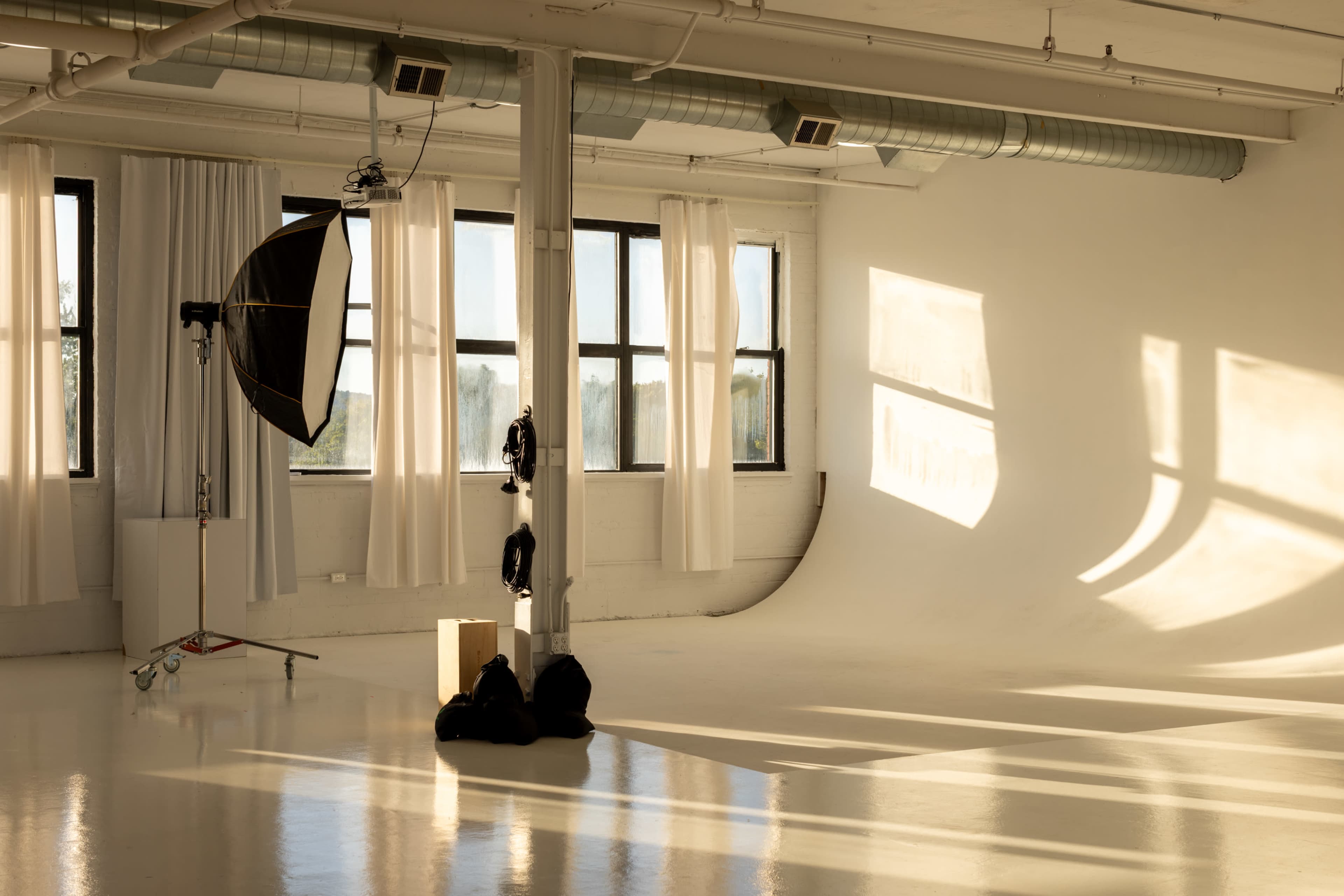 The image shows a photography studio with natural light streaming in through large windows, illuminating a seamless backdrop and various equipment.