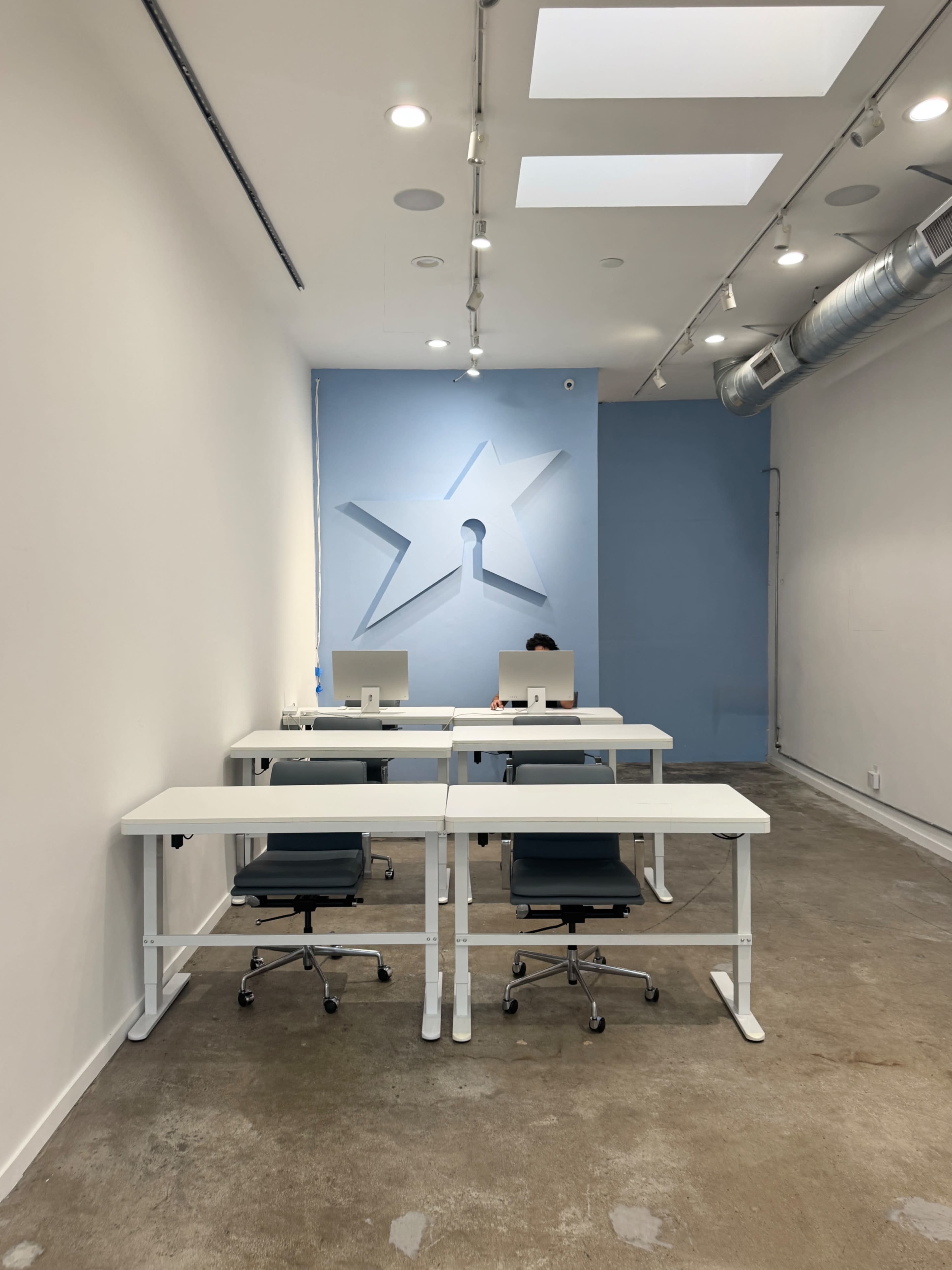 A minimalist office space features two desks with computers and a single person sitting at one desk.