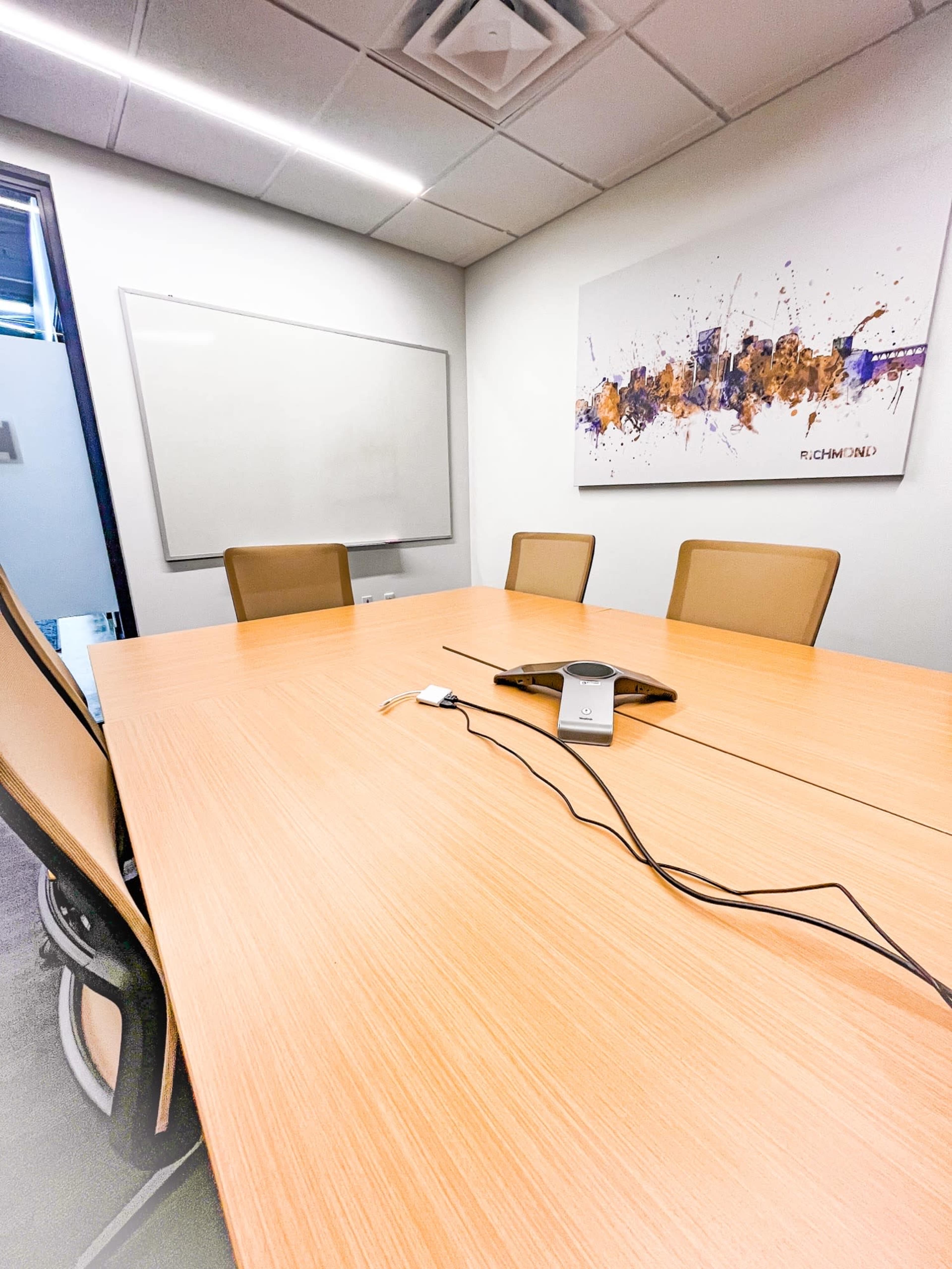 A neatly arranged conference room with a wooden table, four chairs, a whiteboard, and a mounted artwork on the wall.