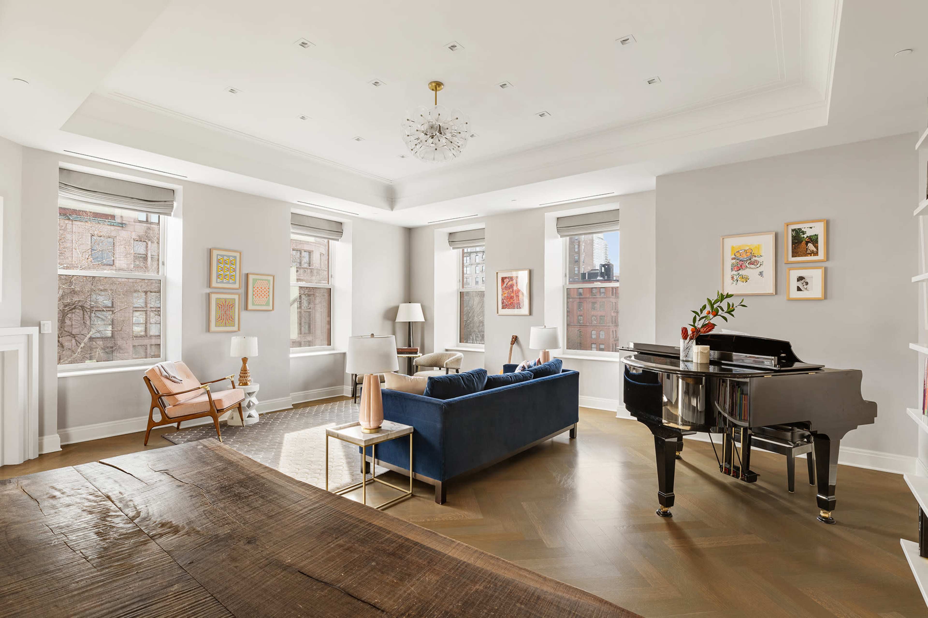 A bright living room features a blue sofa, a black grand piano, and large windows with city views.
