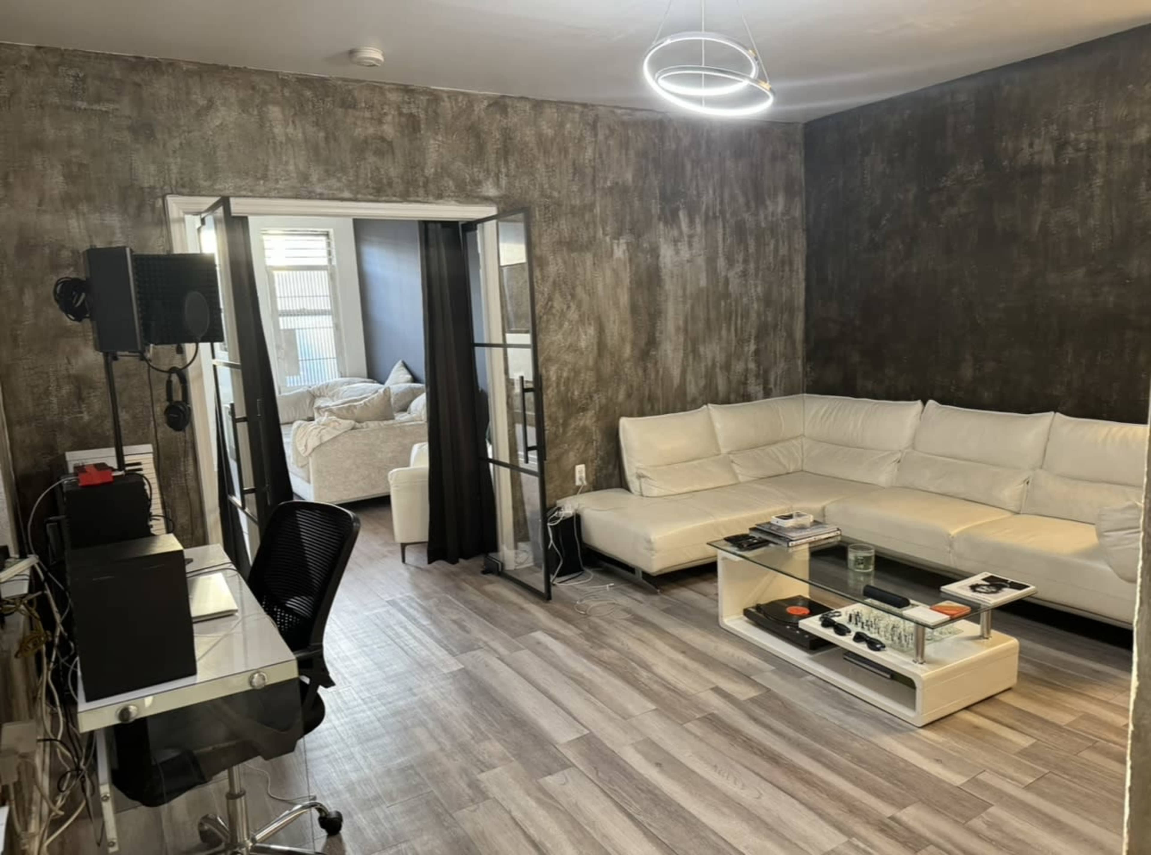 A modern living room with a white sectional sofa, a glass coffee table, and a desk with a computer setup, leading to an adjoining bedroom through glass doors.