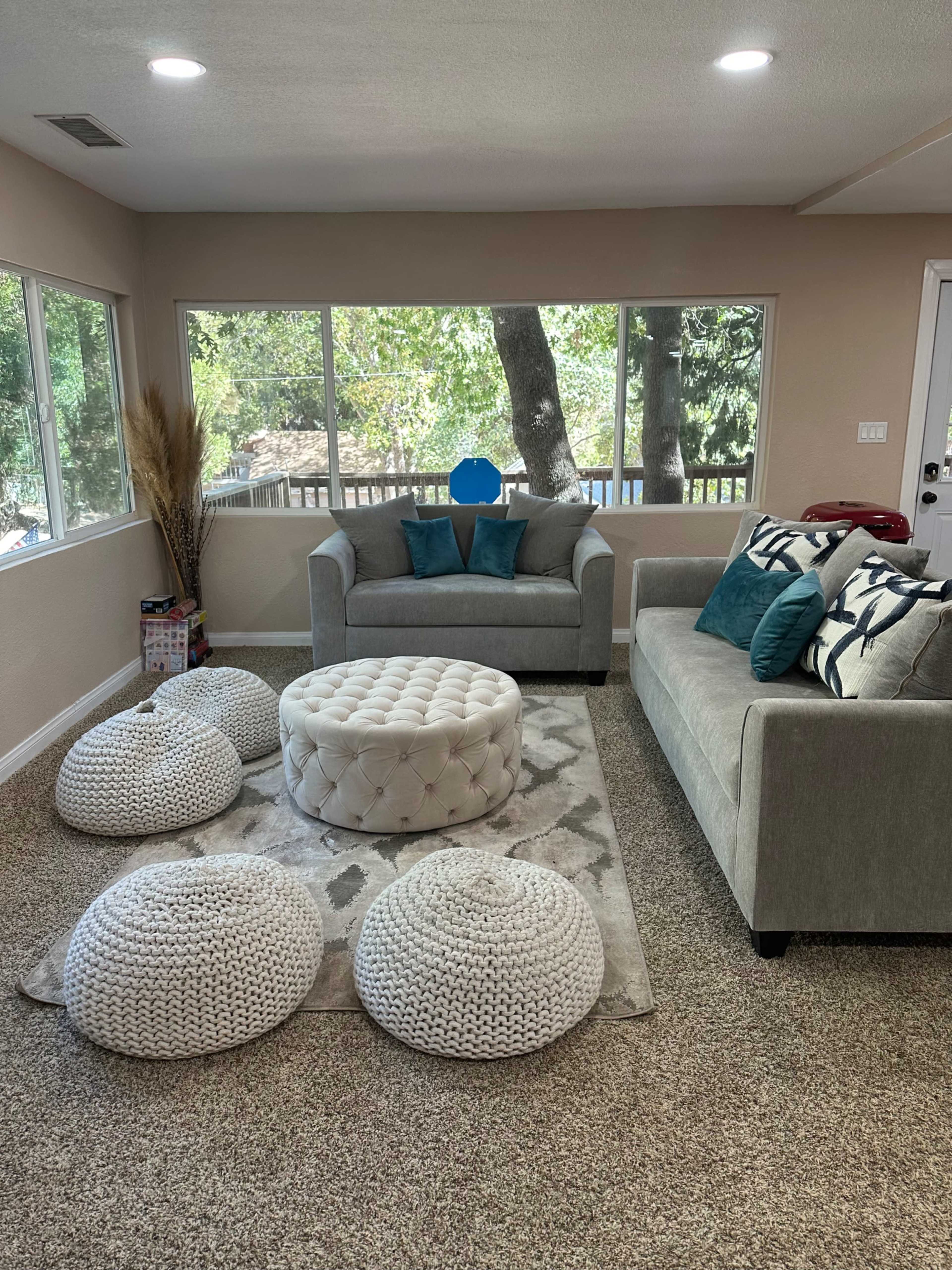 The image shows a light-colored living room with a couch, armchair, tufted round ottoman, and knit poufs, all arranged around a patterned area rug under large windows overlooking greenery.