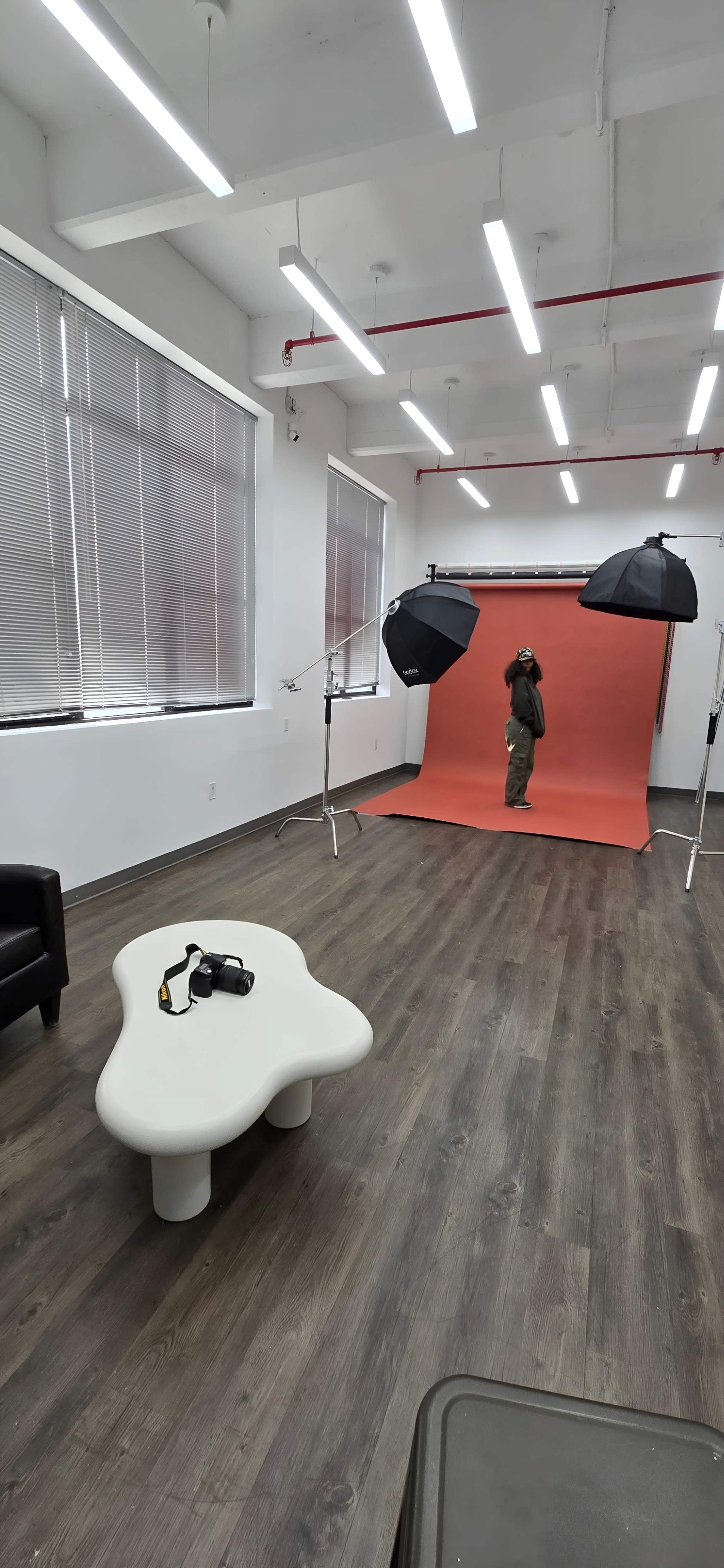 The image shows a photo studio with an orange backdrop, two softbox lights, a white table, and a person standing in the center.