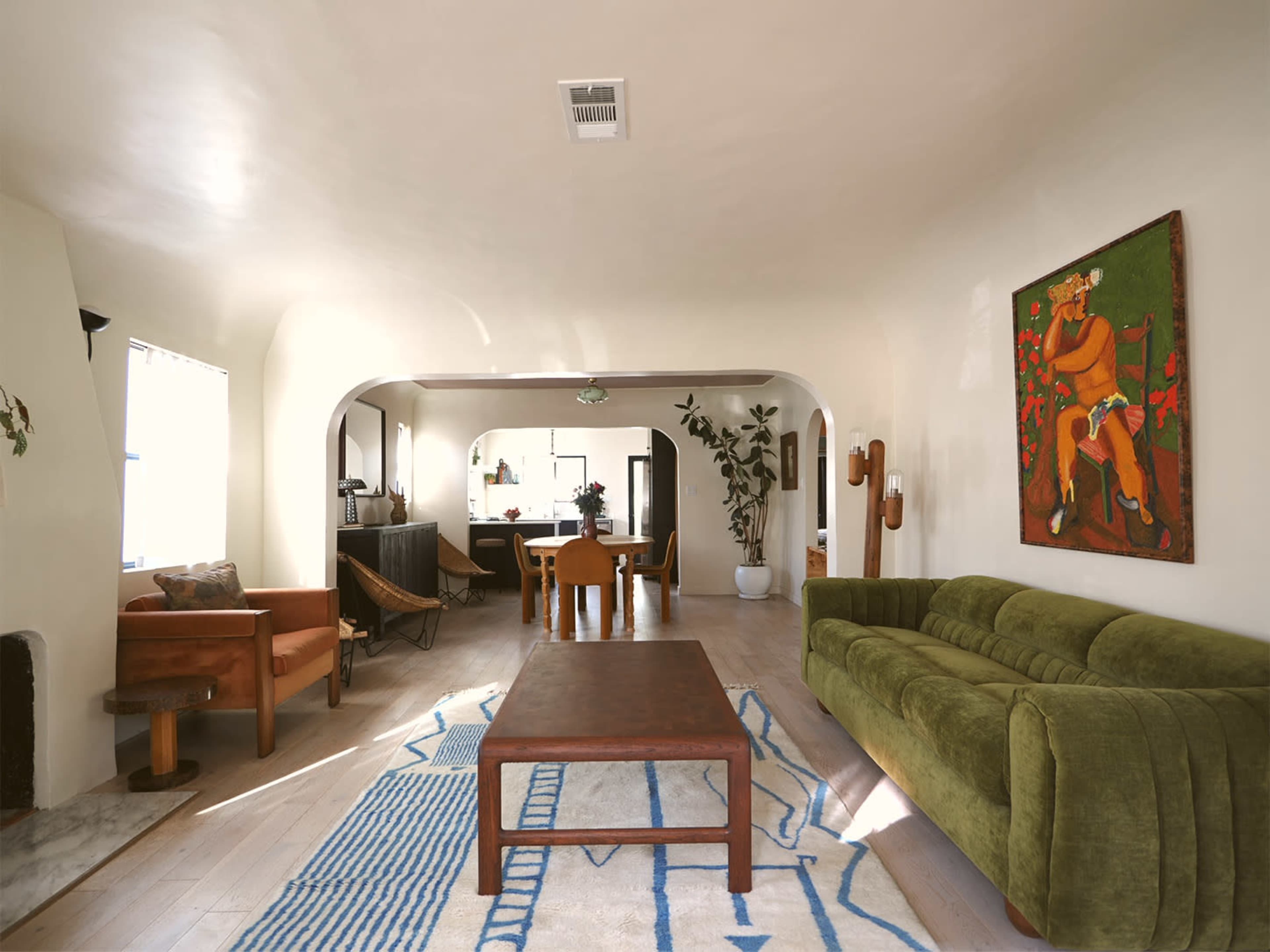 The image shows a spacious living area featuring a green sofa, a wooden coffee table, and a vibrant artwork on the wall, with natural light illuminating the room.