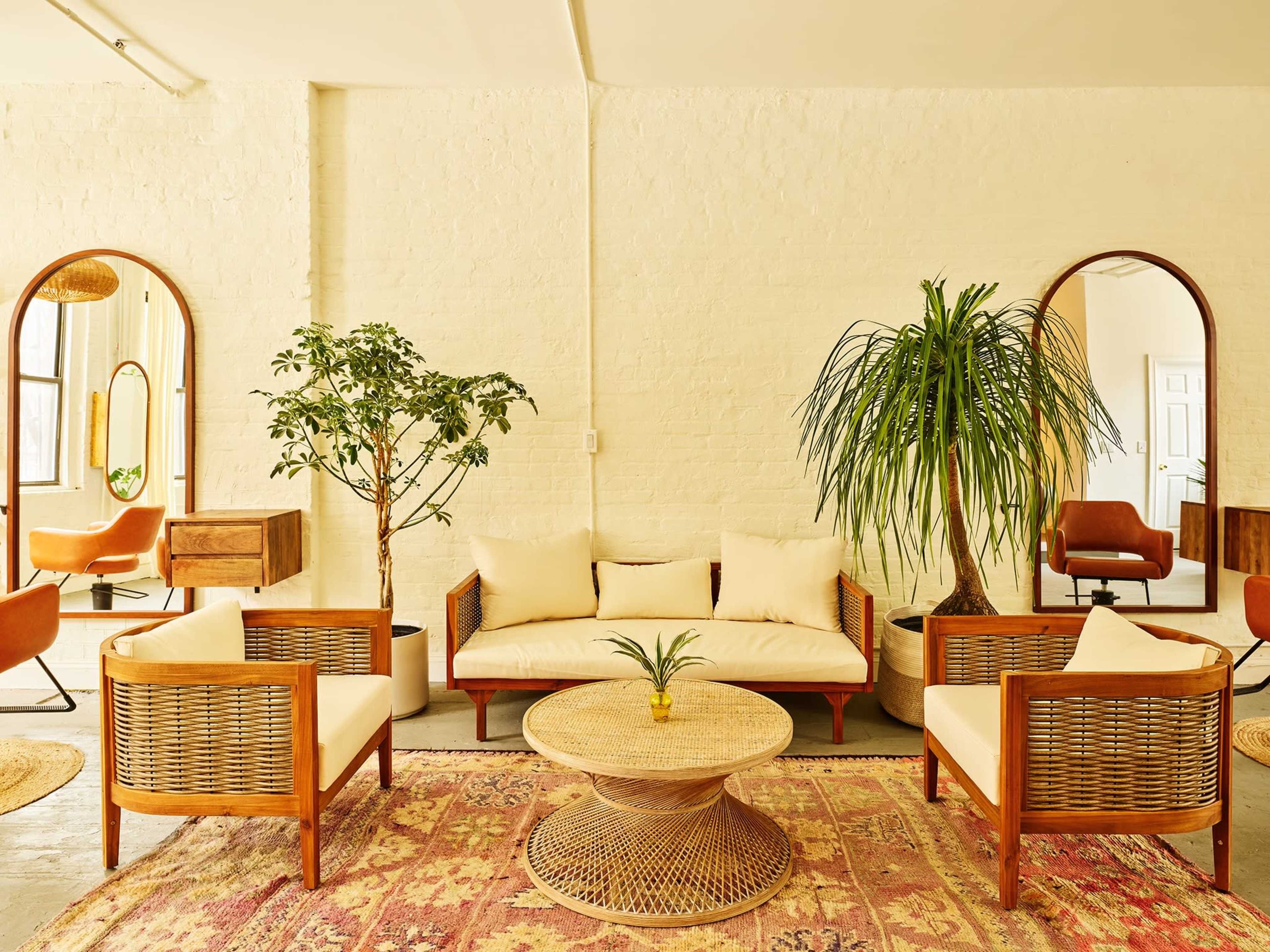 The image shows a bright, airy lounge area with rattan furniture, a round coffee table, and various plants, set against a textured white brick wall.