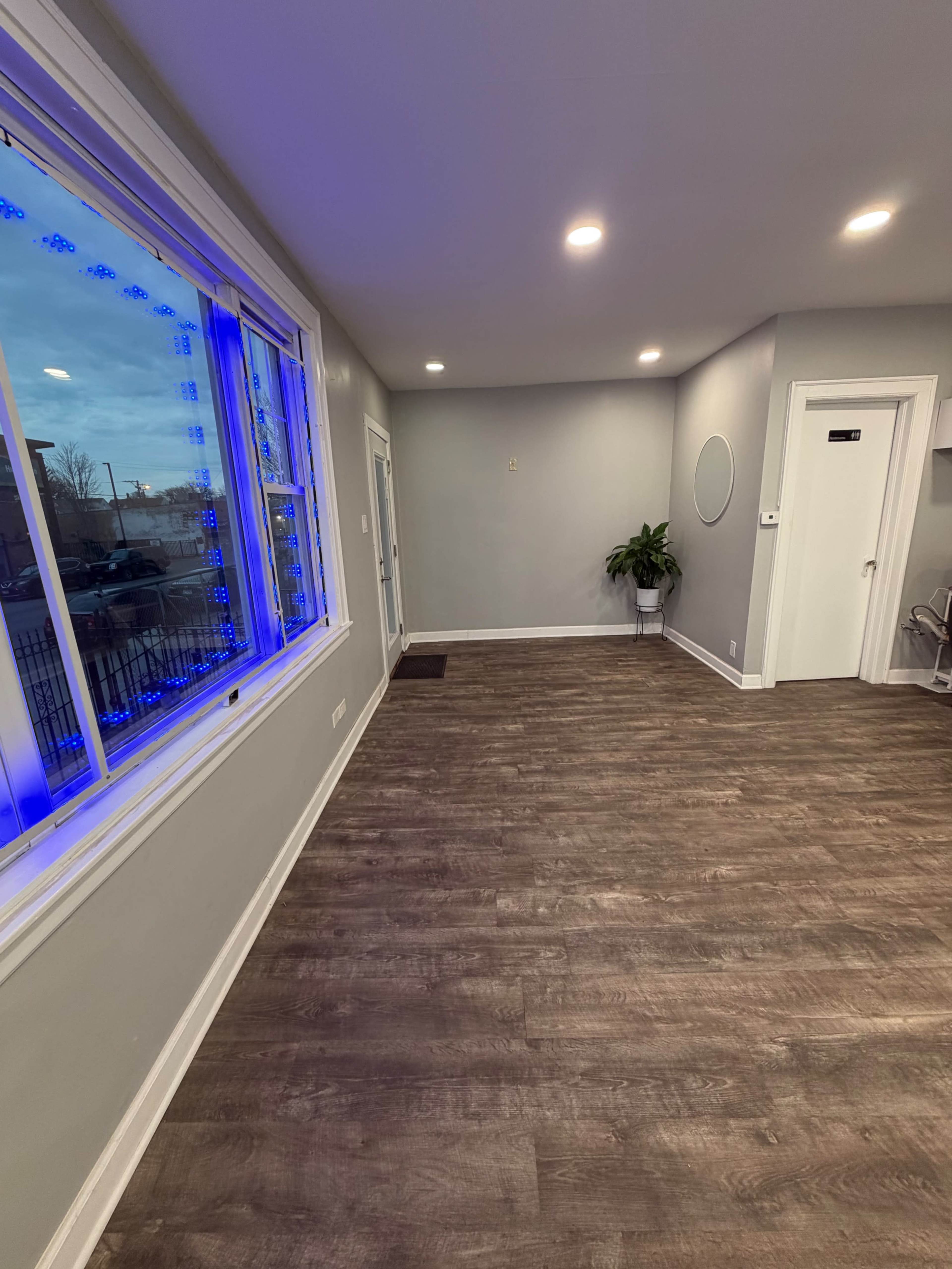 The image shows a spacious room with wooden flooring, a large window with blue lighting, and minimal furniture.