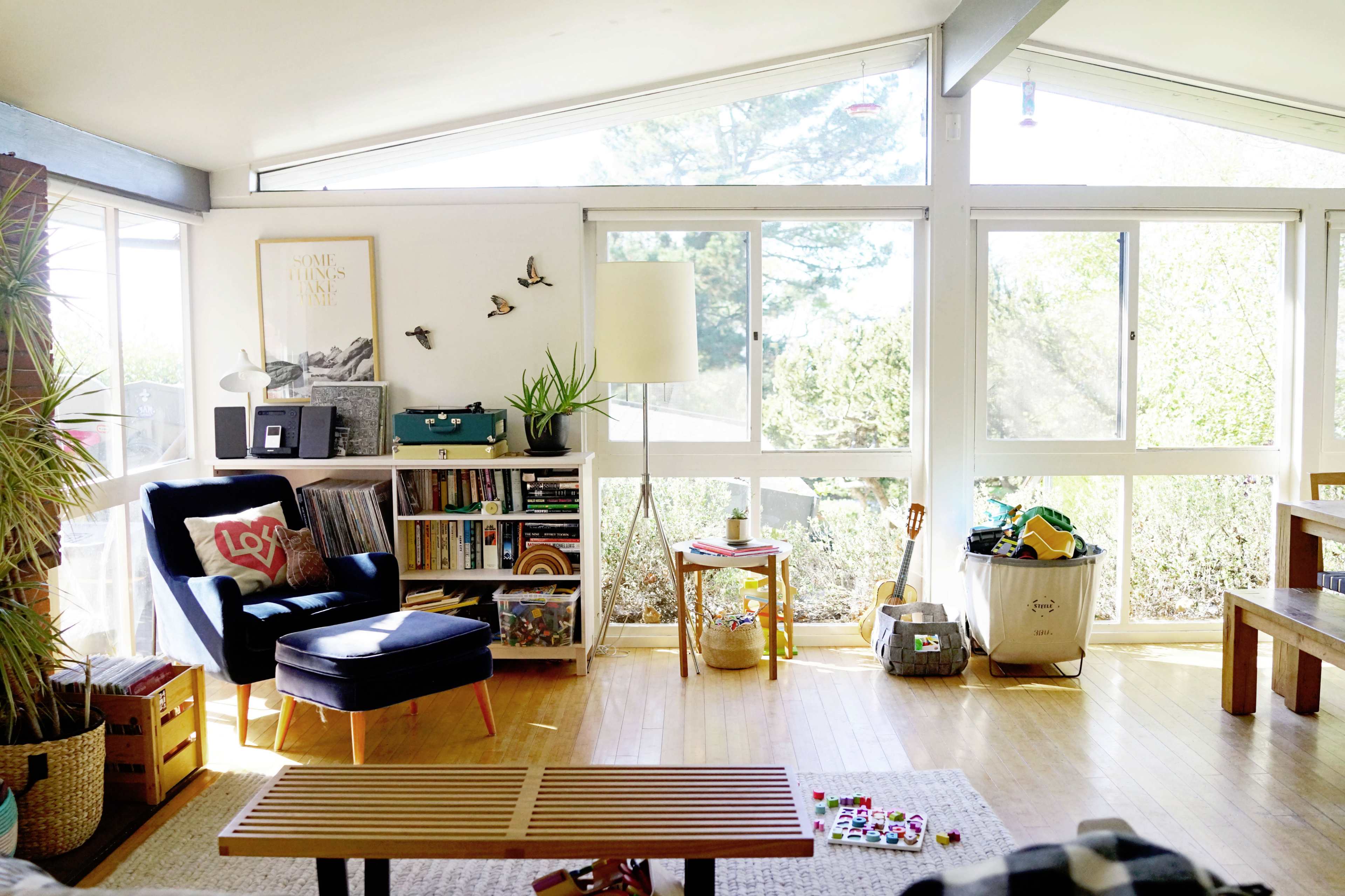 The living room features a large window with natural light, a blue armchair, a wooden coffee table, and various plants and decor items.