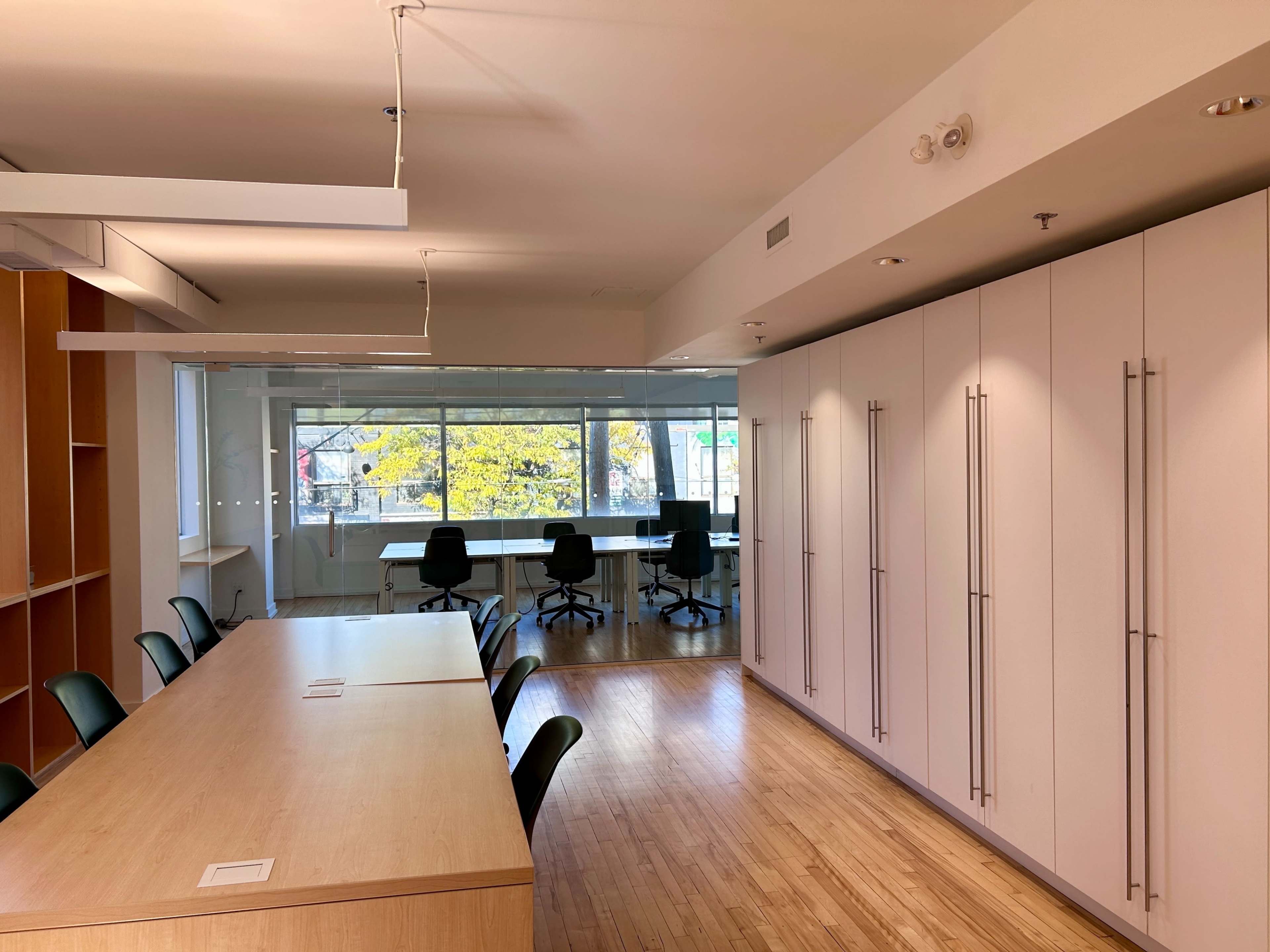 The image shows a modern office space featuring a long wooden table with chairs, a wall of cabinets, and a conference area with large windows allowing natural light to enter.