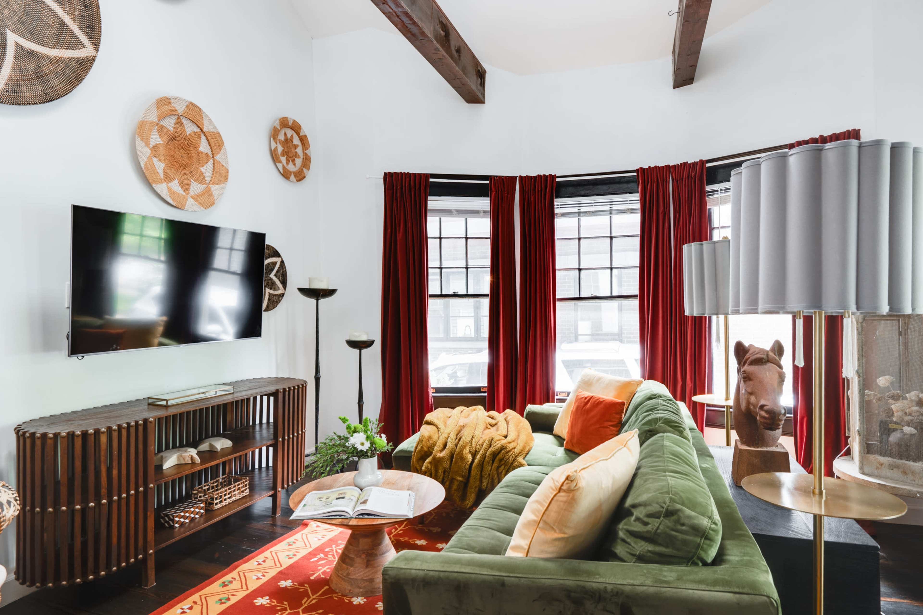 The image shows a cozy living room featuring a green velvet sofa, a large flat-screen television mounted on the wall, and decorative baskets hanging above the windows.