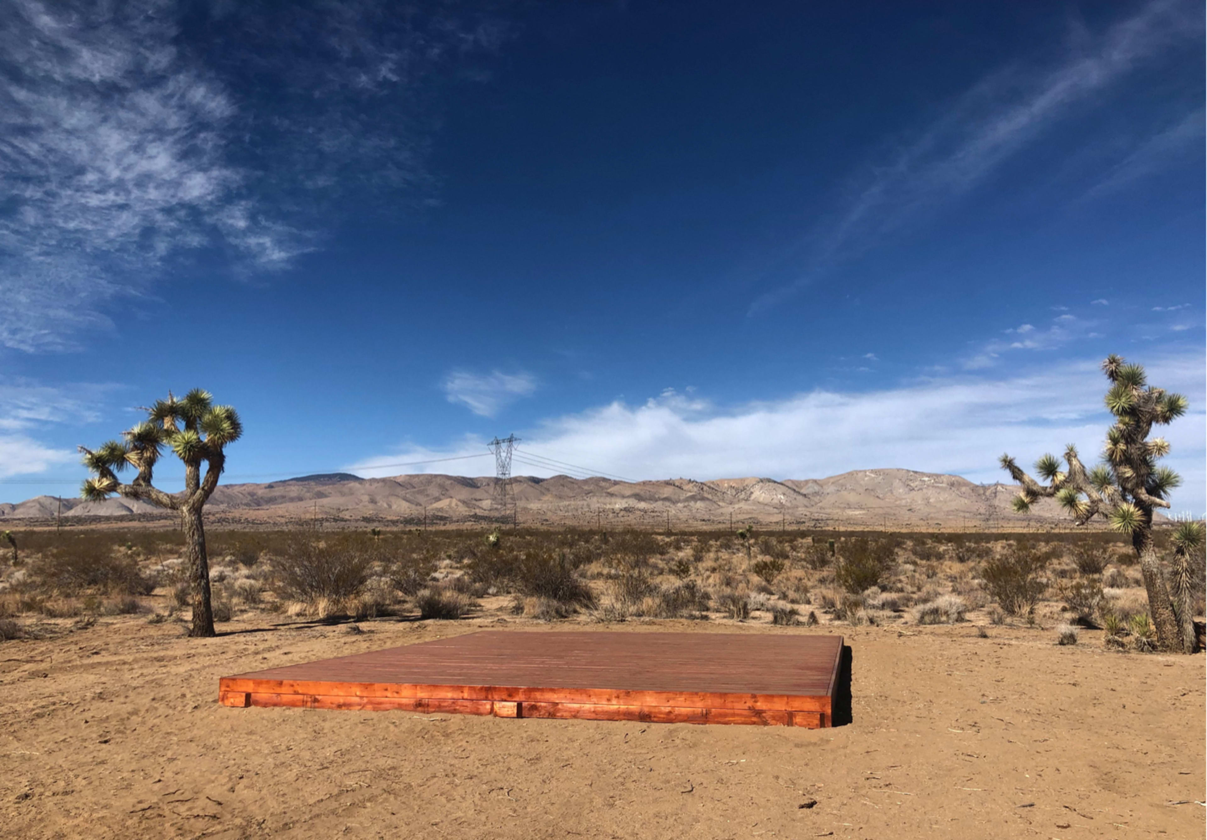 A flat wooden platform is set on dry ground surrounded by sparse desert vegetation and distant mountains under a blue sky.