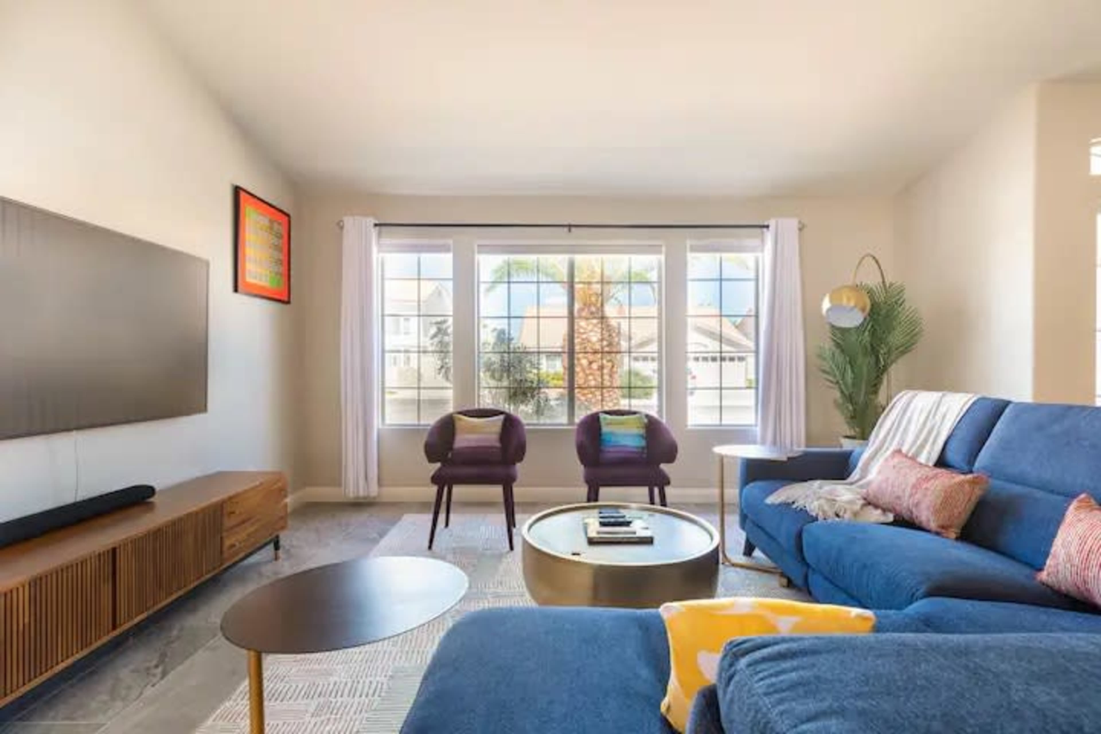 A bright living room features a blue sectional sofa, a round coffee table, and two purple armchairs near large windows with views of palm trees.