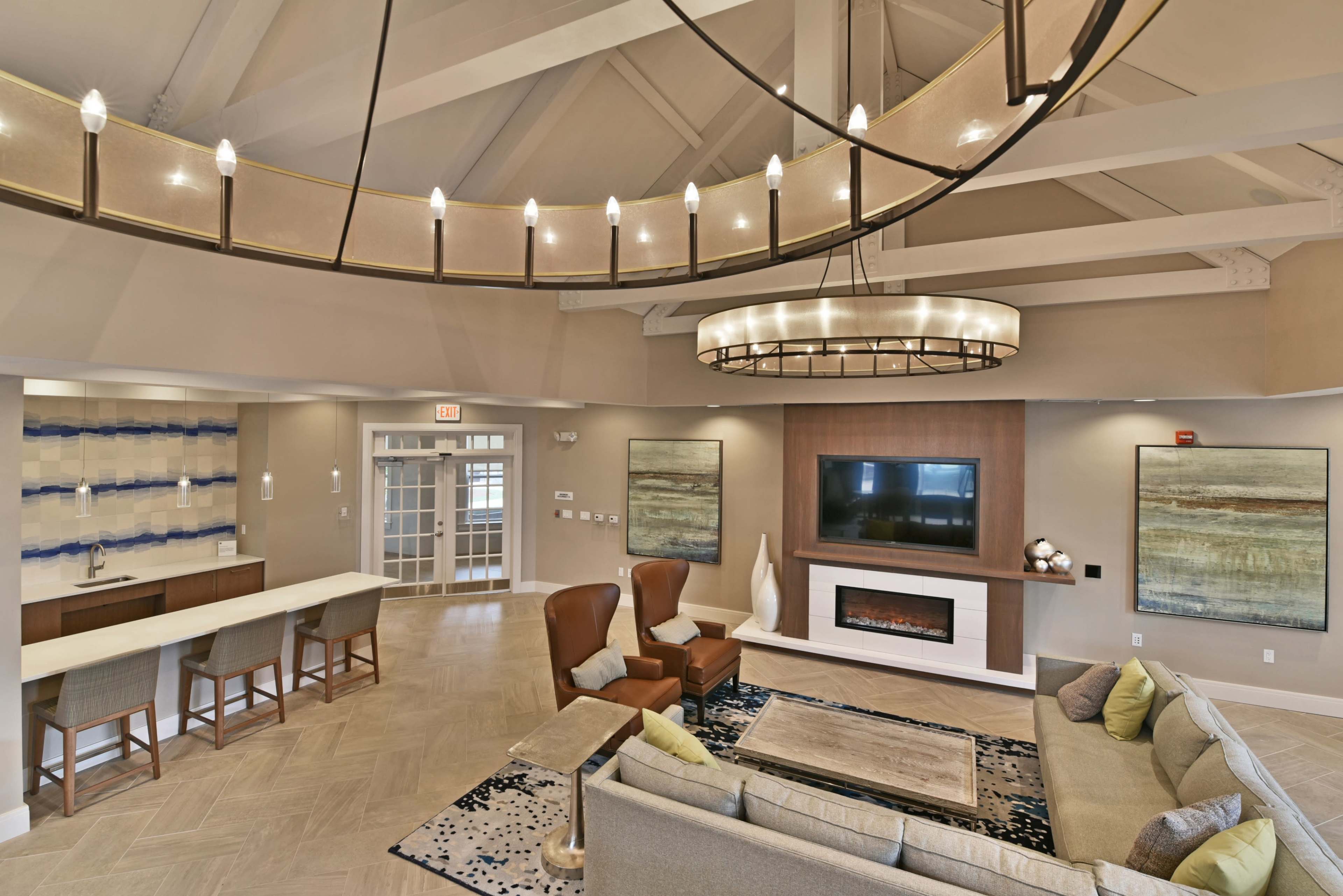 The image shows a modern living room with a high ceiling, featuring a comfortable seating area, a wall-mounted television, and a decorative fireplace.