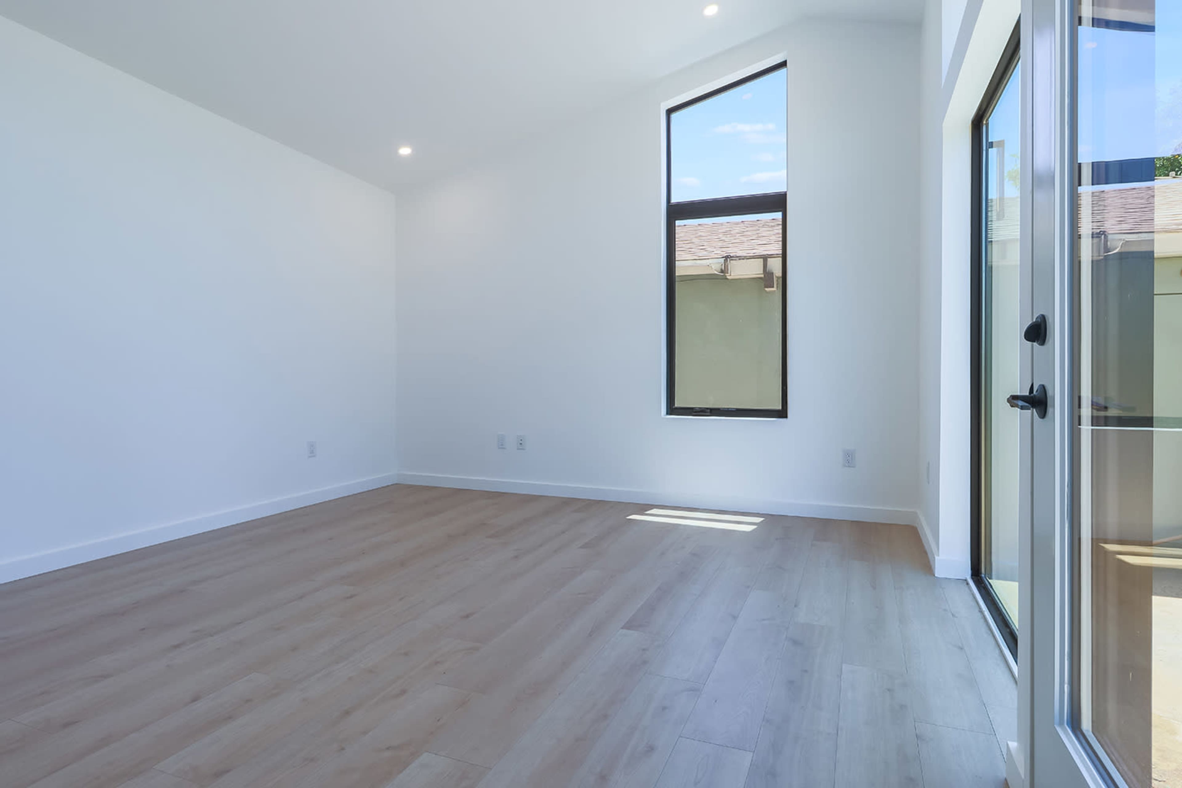An empty room with large windows allowing natural light and light-colored wooden flooring.