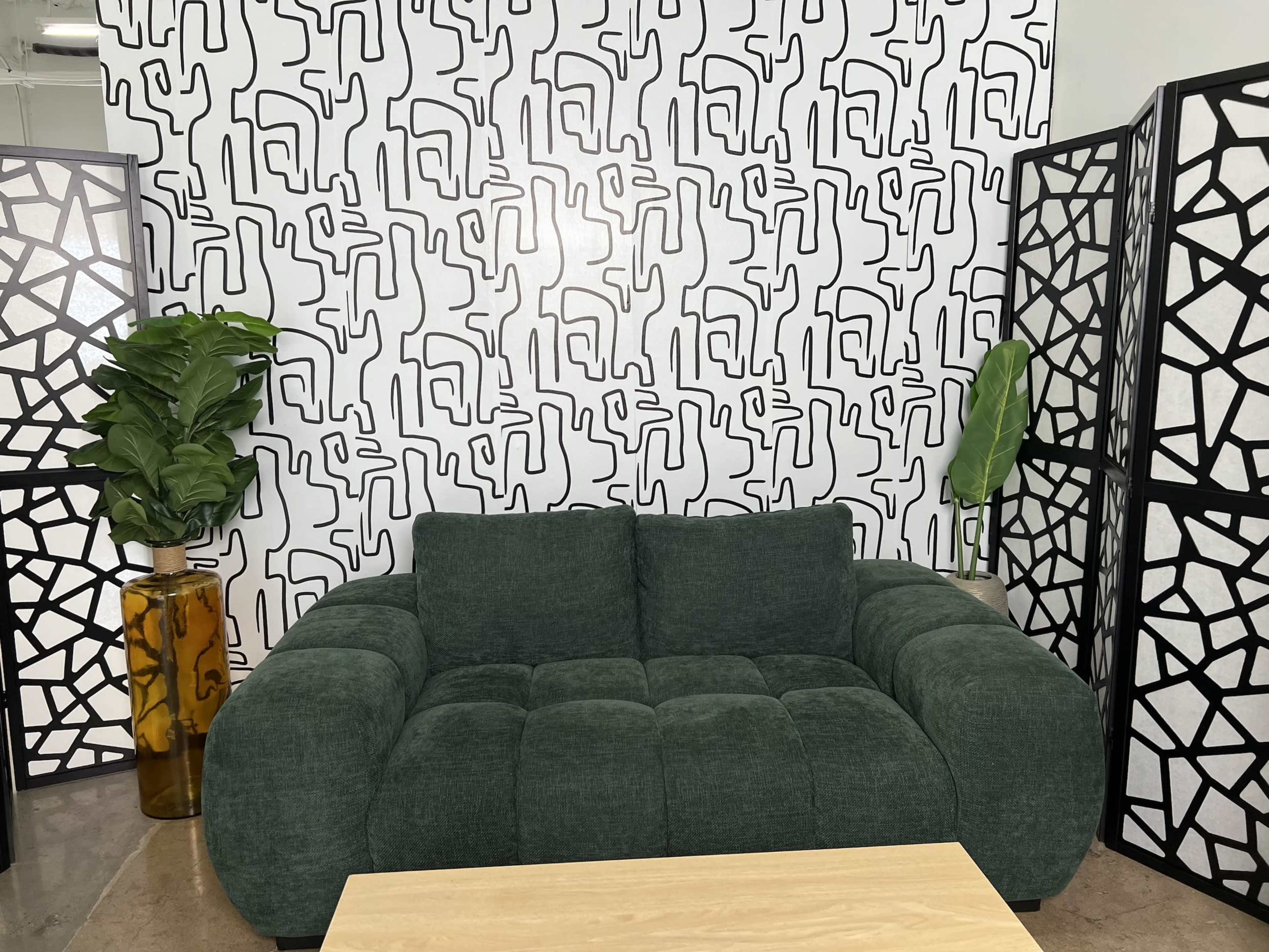 The image features a green couch in front of a patterned black and white wall, accompanied by a tall plant in a yellow vase and a wooden table.