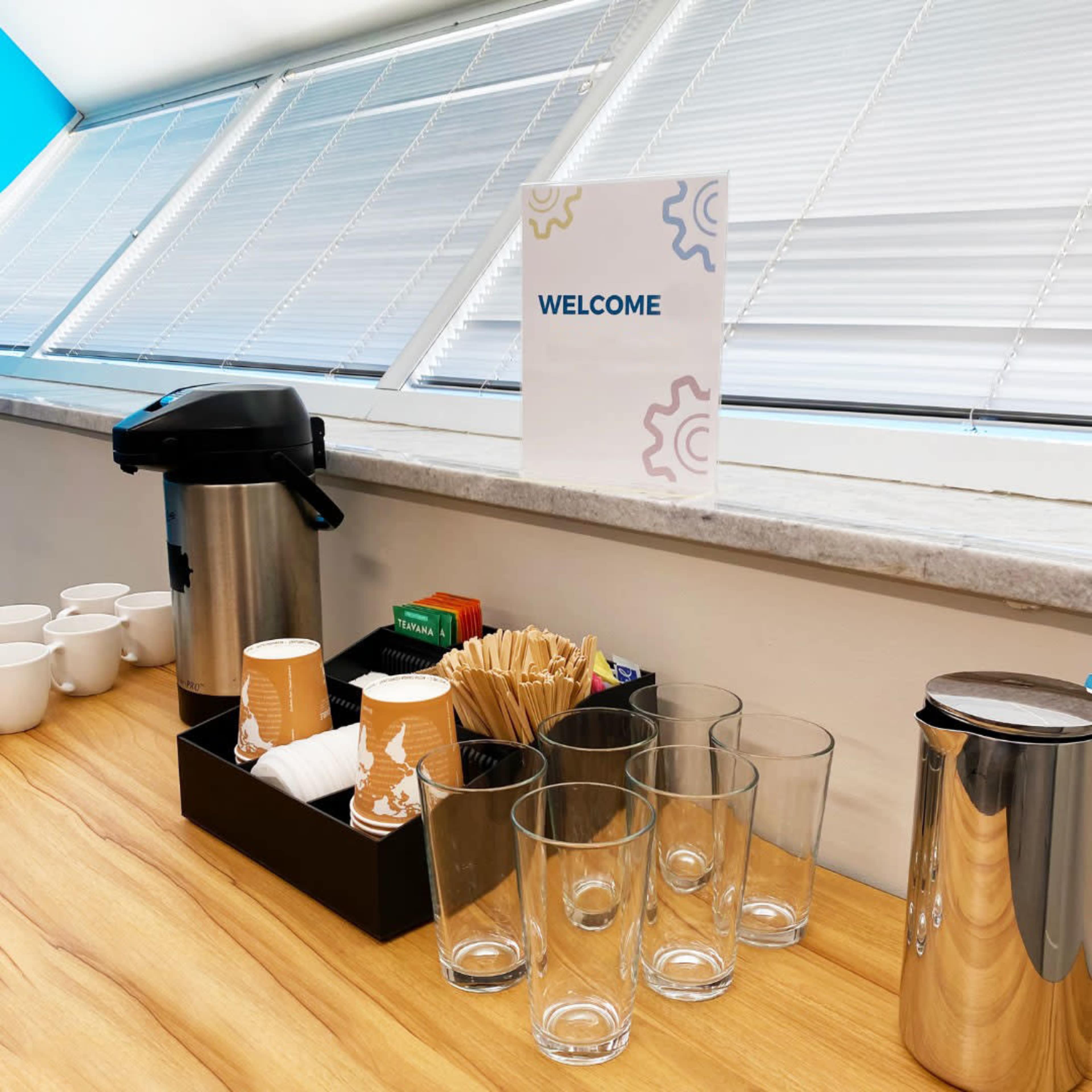 A coffee station is set up on a wooden countertop with cups, glasses, a thermos, utensils, and a welcome sign.