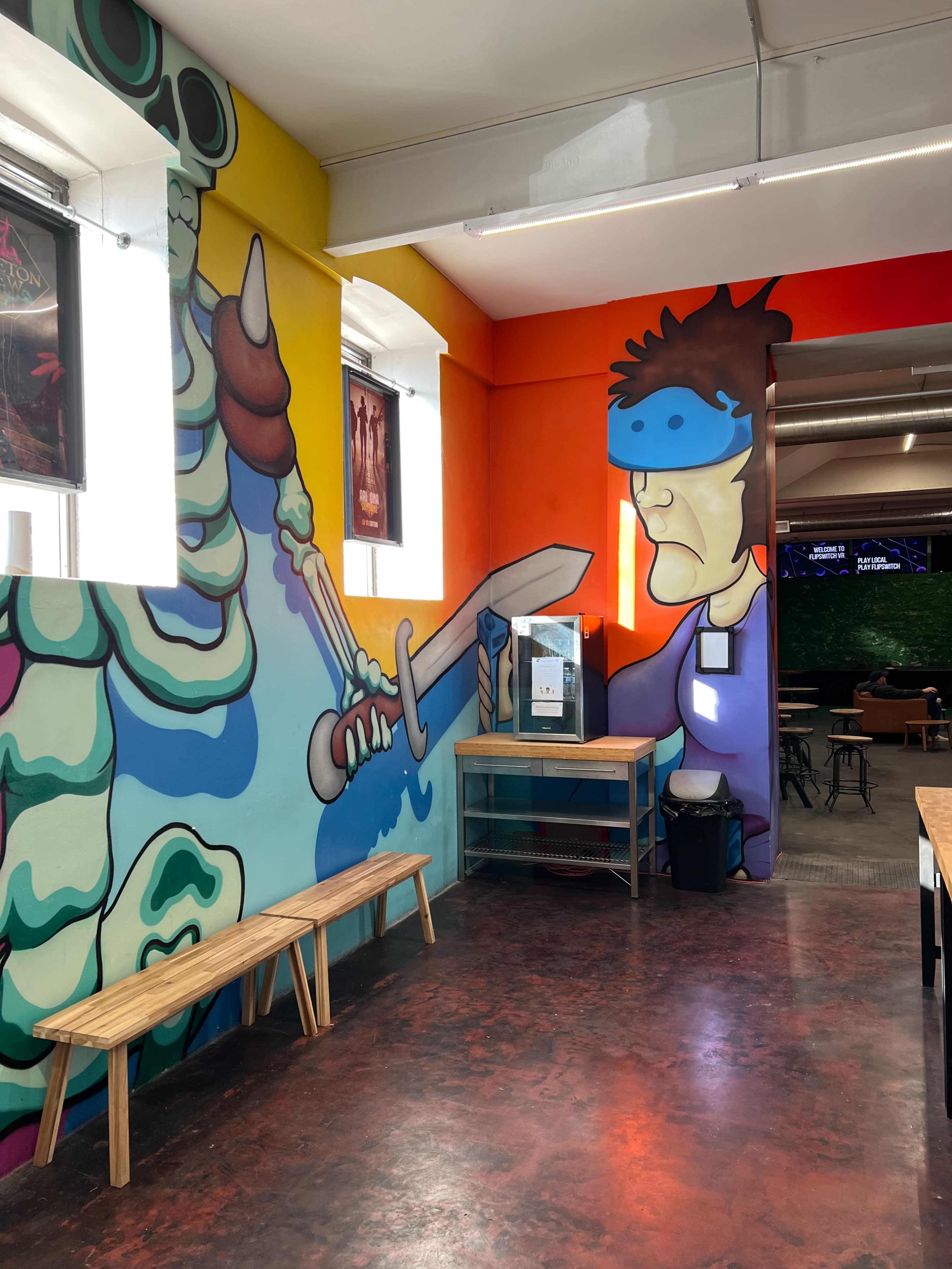 A brightly colored mural featuring a character with a sword, interacting with a stylized creature, alongside wooden benches and a table in a modern cafe setting.
