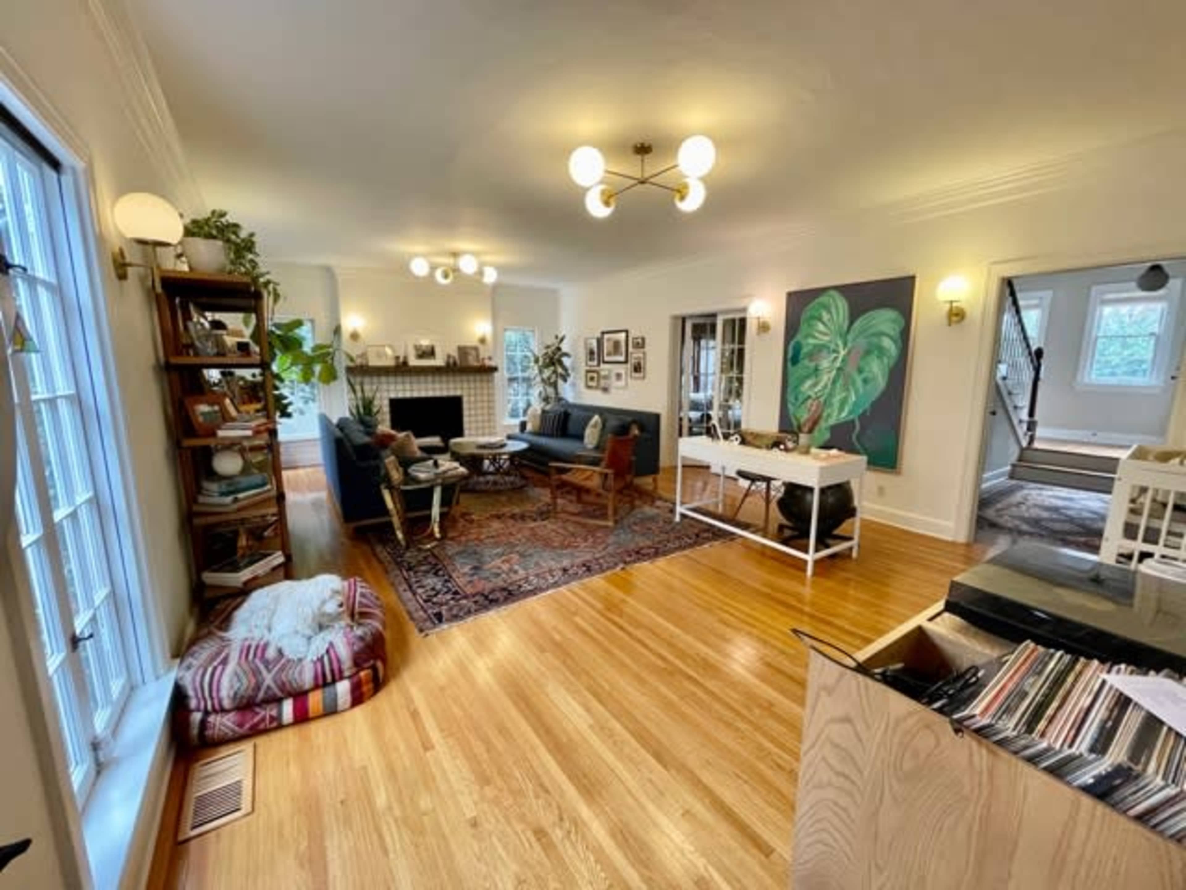 A spacious living room featuring hardwood floors, a fireplace, two sofas, a coffee table, and a bookshelf, all surrounded by plants and artwork.