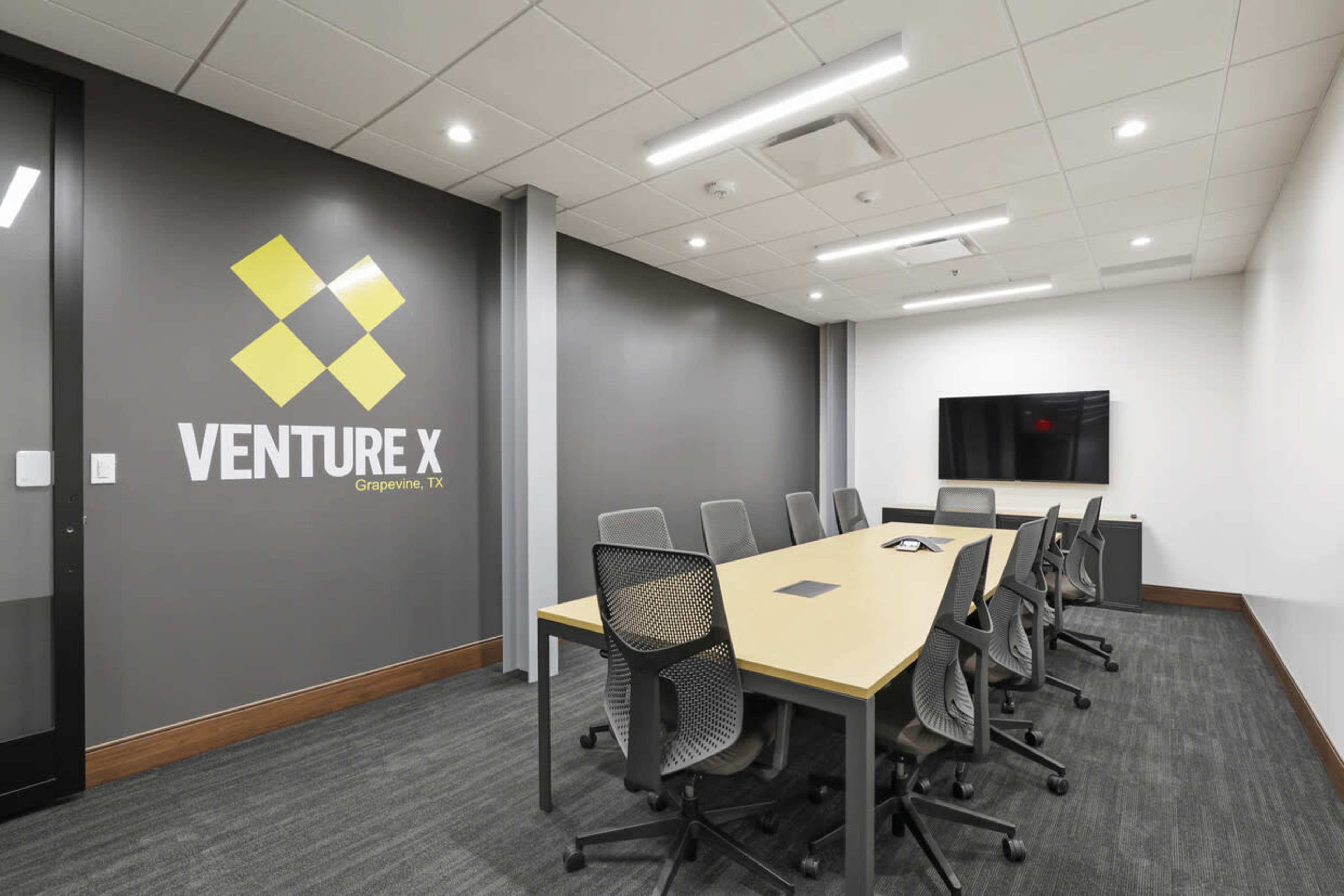 A modern meeting room with a long table, ergonomic chairs, a large screen, and a wall featuring the "VENTURE X" logo.