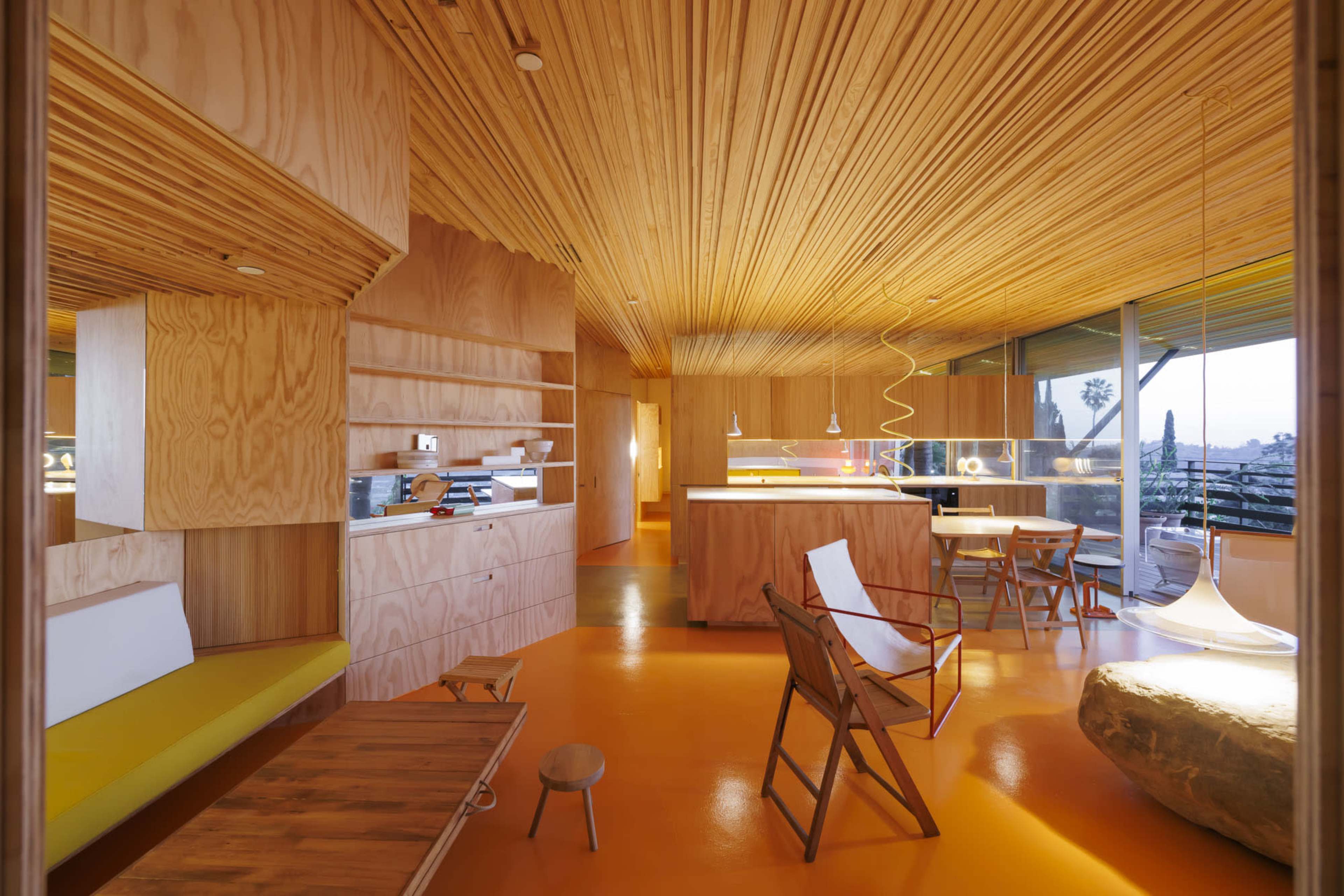 The image shows a modern interior space with wooden walls and ceiling, featuring an open layout that includes a kitchen area, seating, and large windows.
