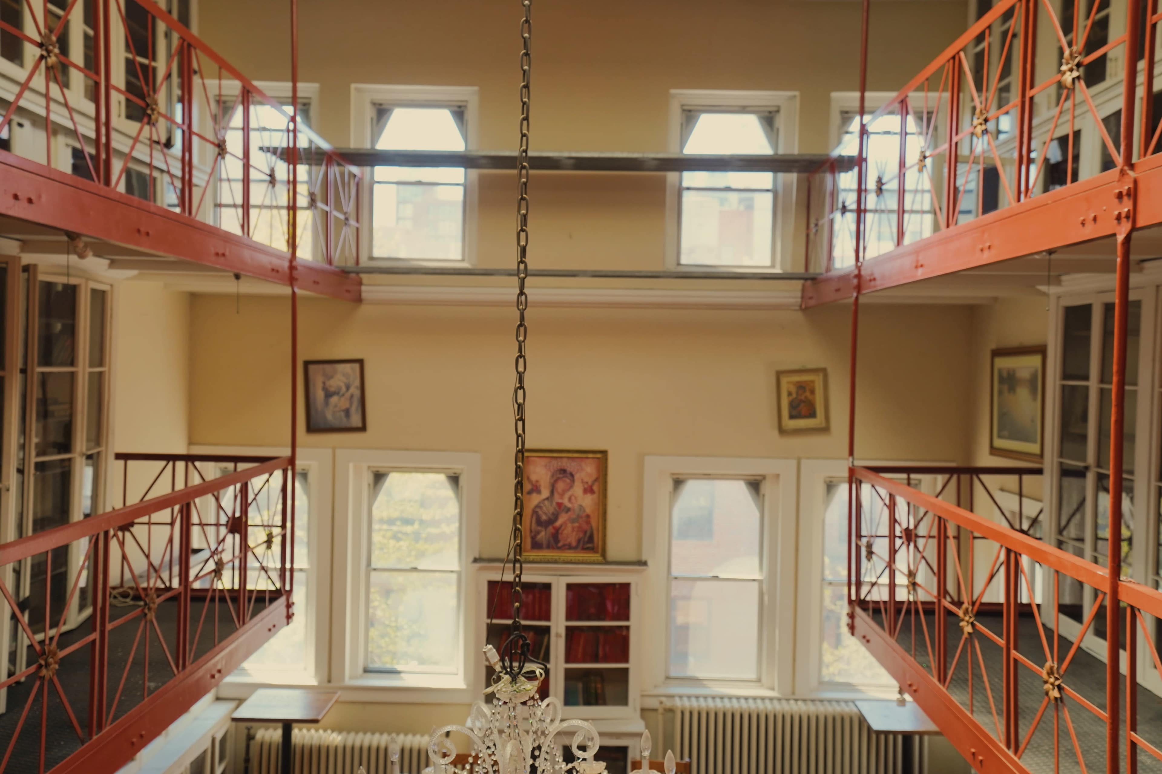 A spacious library with two levels of balcony-style shelves, featuring artwork on the walls and large windows allowing natural light.