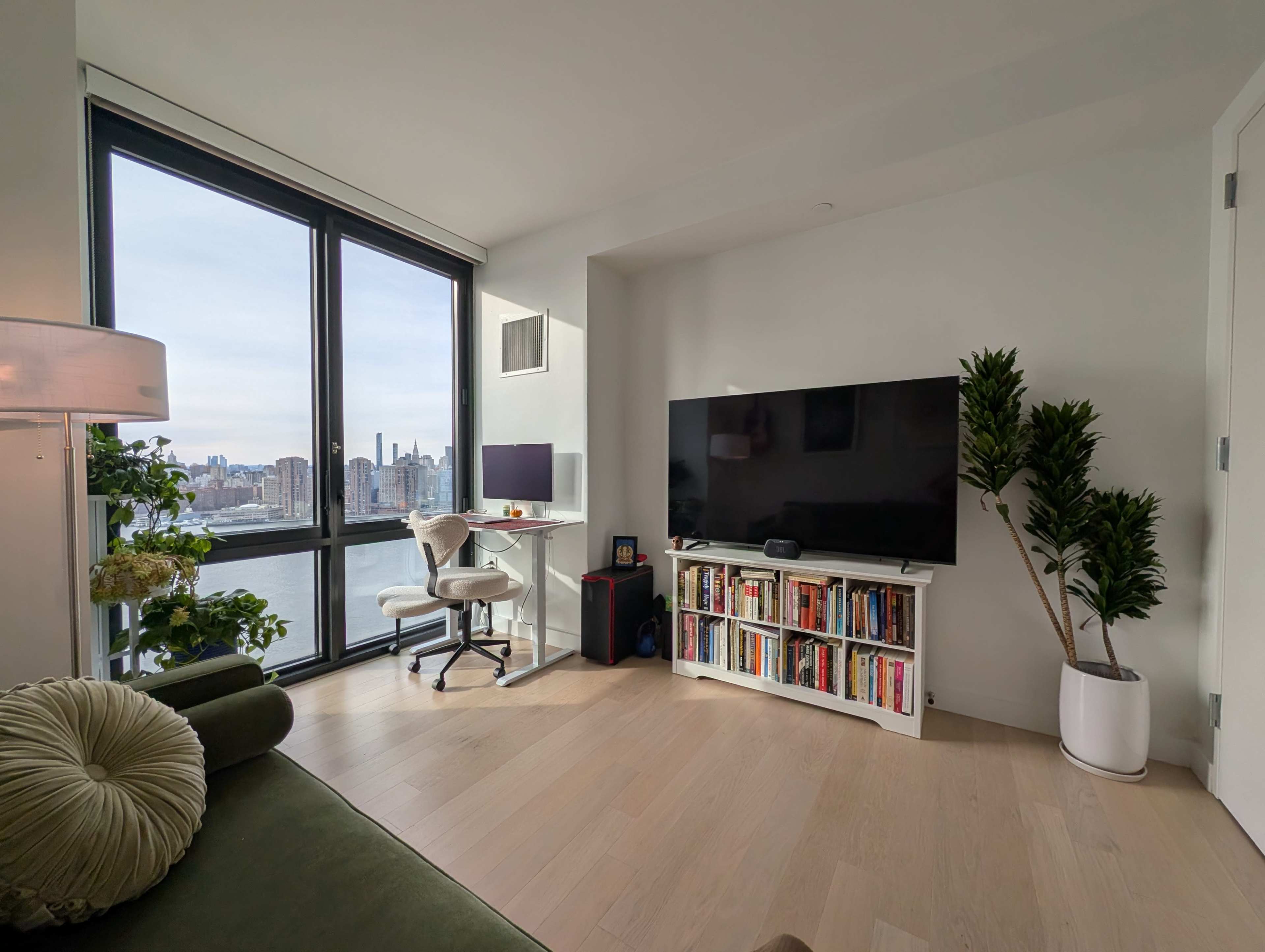 The image shows a modern living room with a large window, a television on a white unit, a desk with a computer, and a bookshelf filled with books.