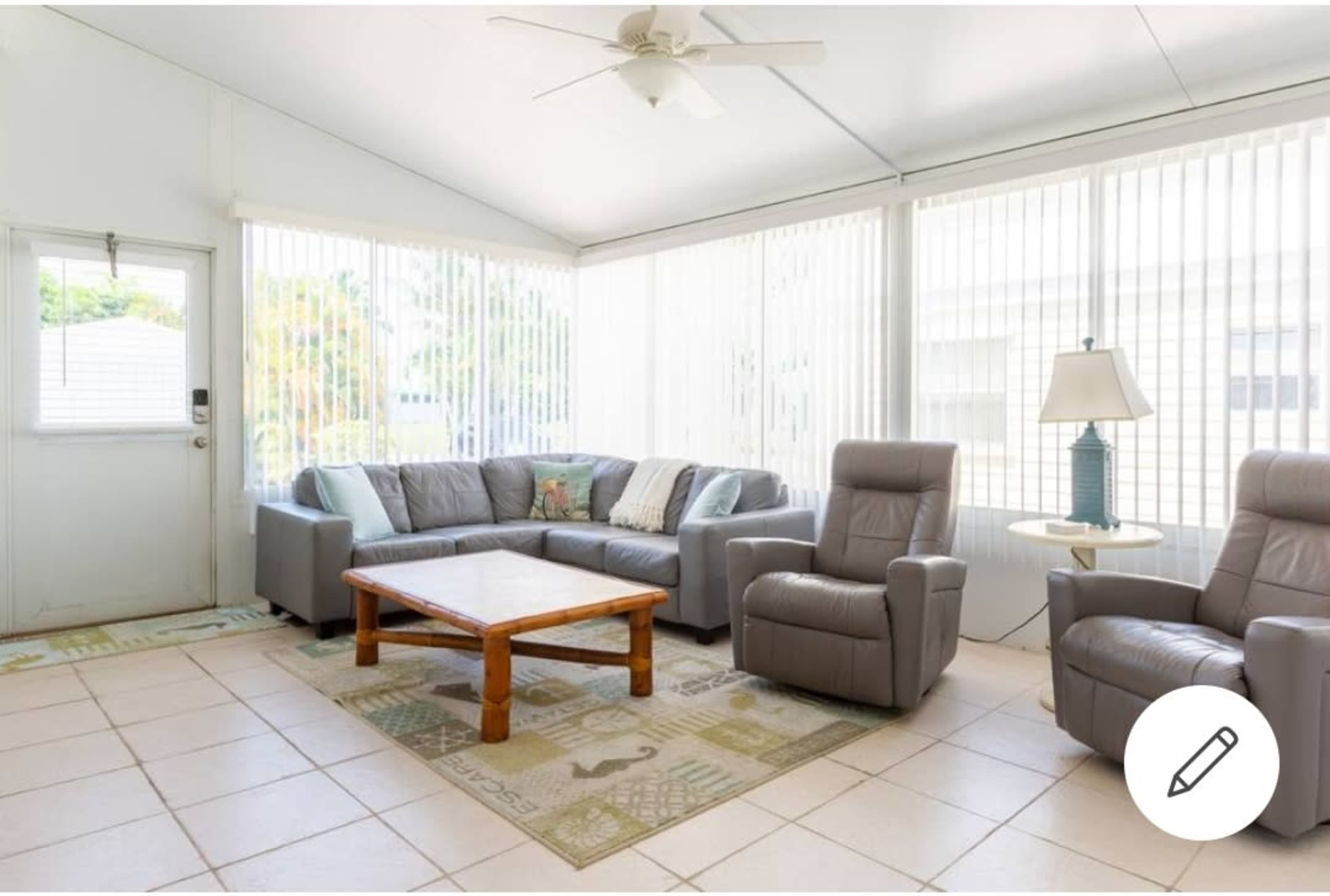 A bright sunroom with beige tiled floors, a gray sectional sofa, two recliners, a wooden coffee table, and large windows covered with vertical blinds.