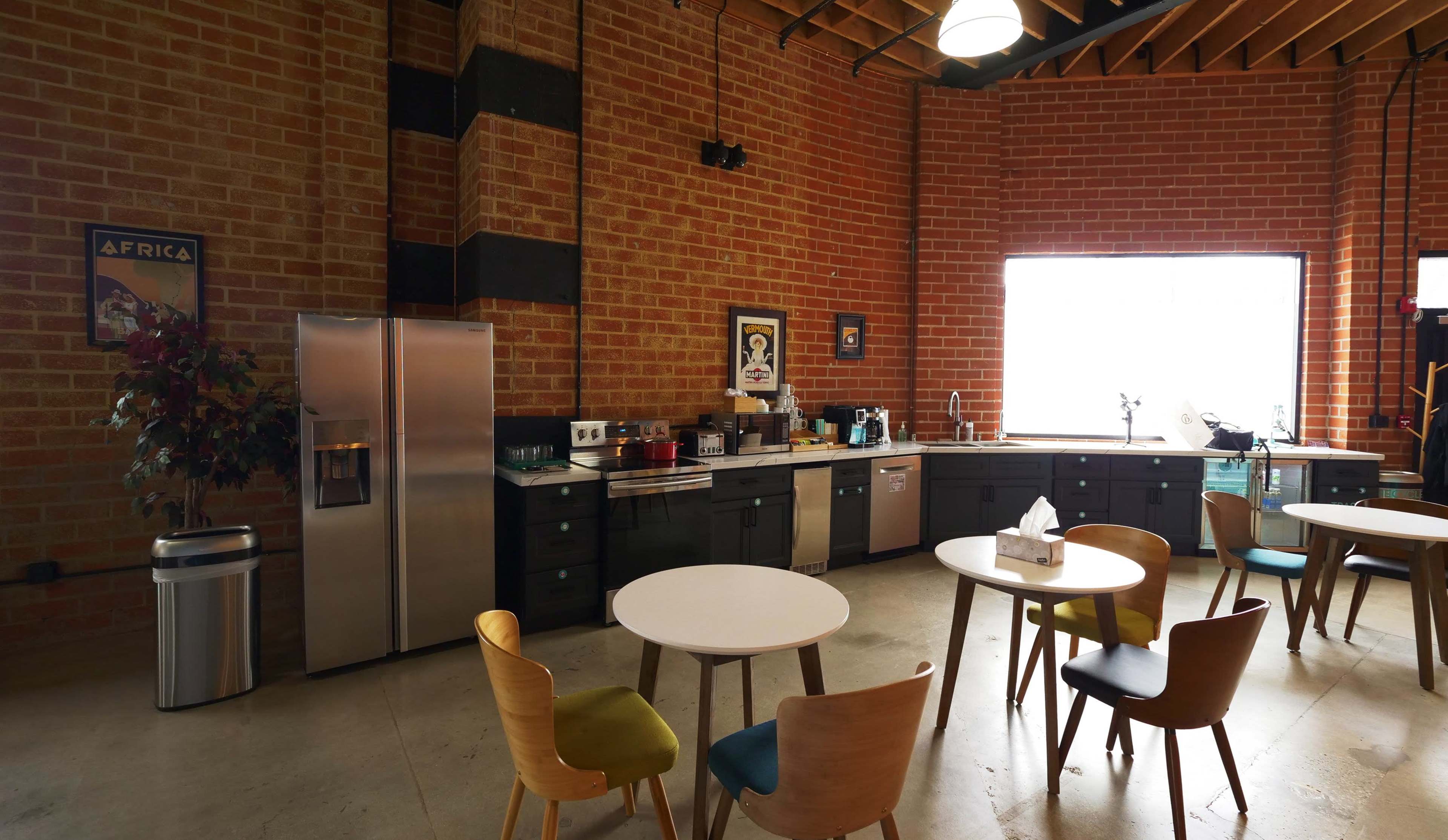 The image shows a modern kitchen area featuring stainless steel appliances, black cabinets, a large window, and several tables with colorful chairs.