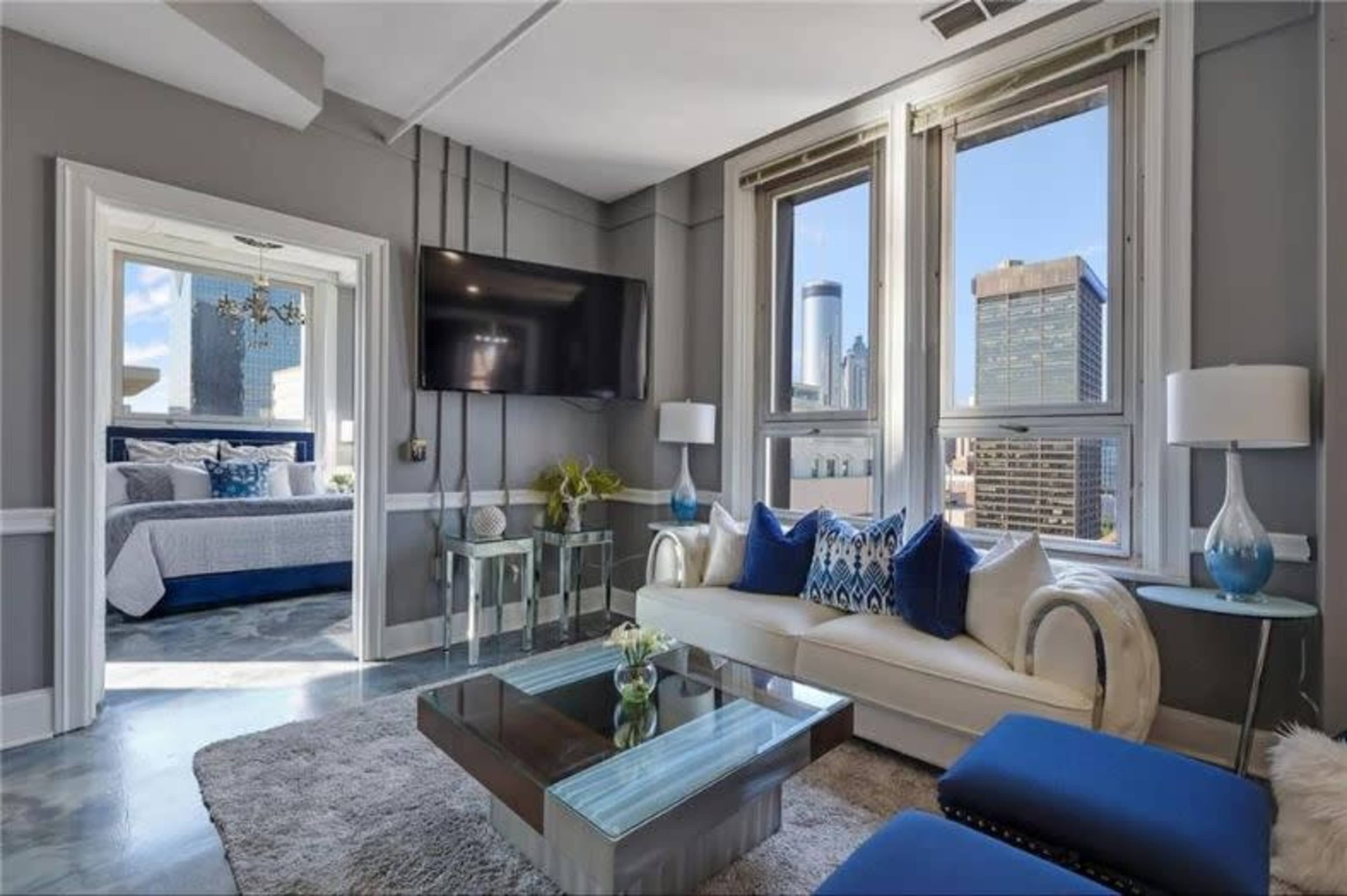 The image shows a modern living room with a white sofa and blue accent pillows, large windows showcasing a city skyline, and a glass coffee table.