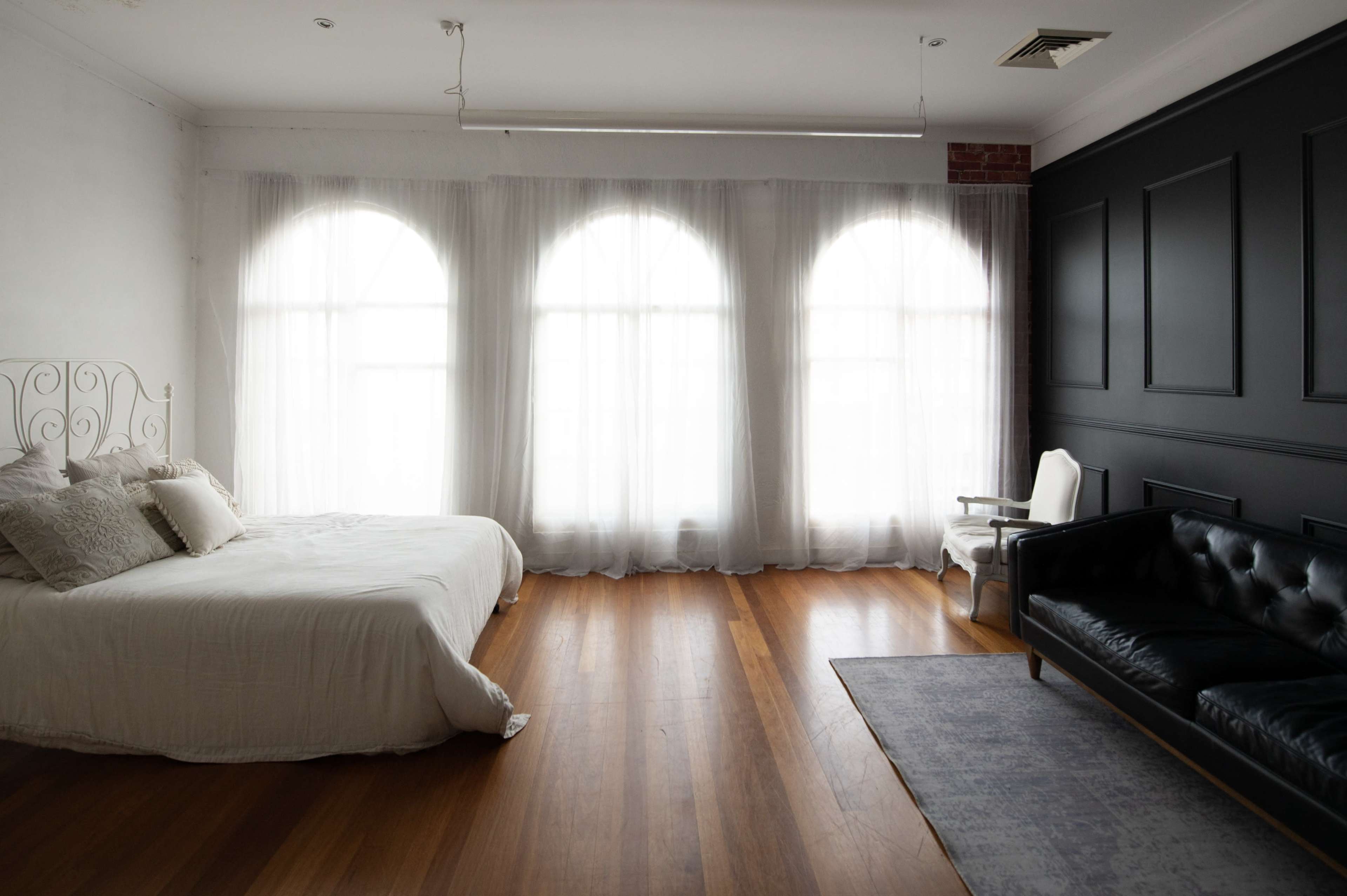The image shows a spacious bedroom with a bed, a black sofa, and large windows covered by sheer curtains.
