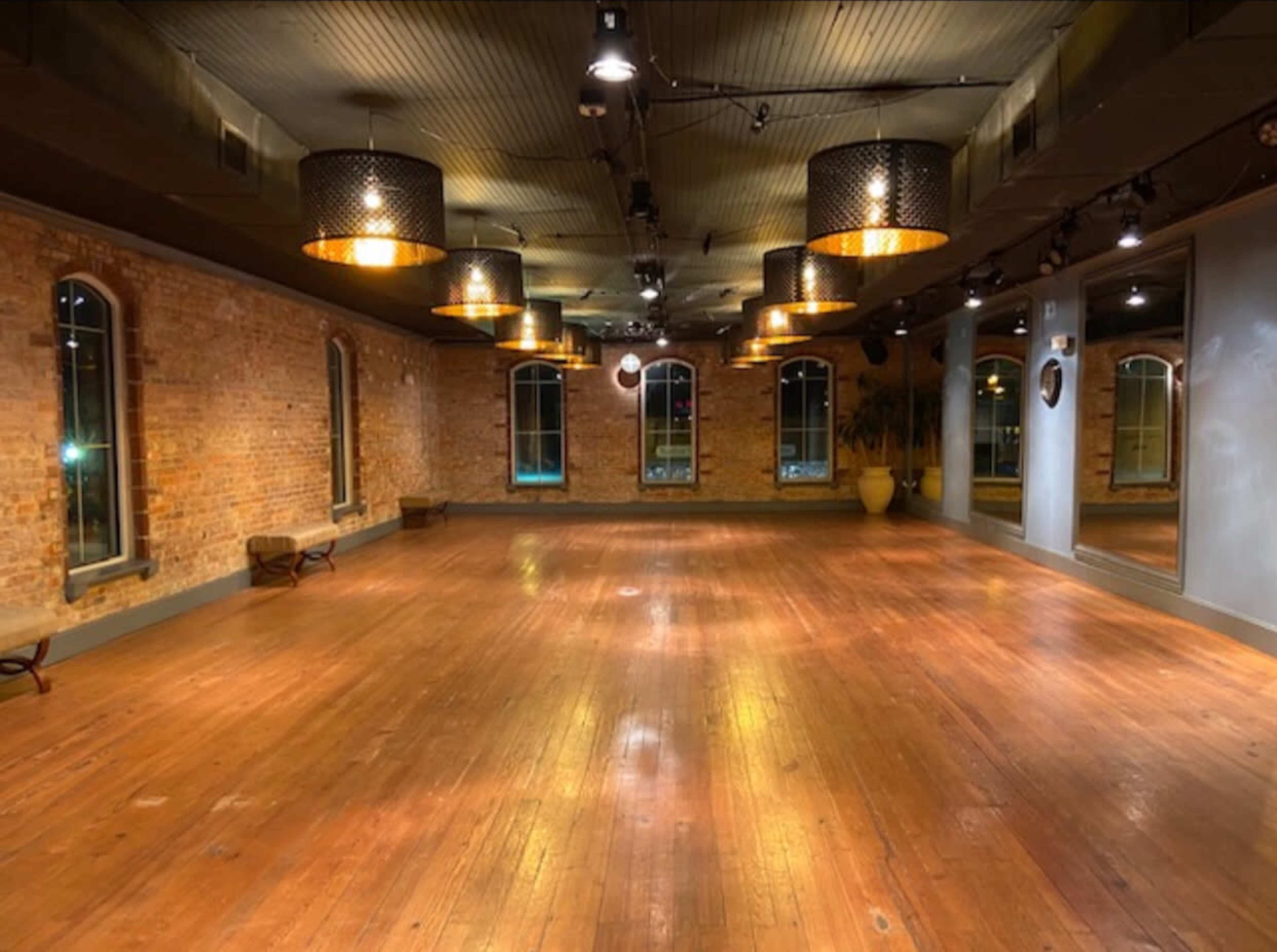A spacious, empty room with wooden floors and exposed brick walls is illuminated by pendant lights hanging from the ceiling.