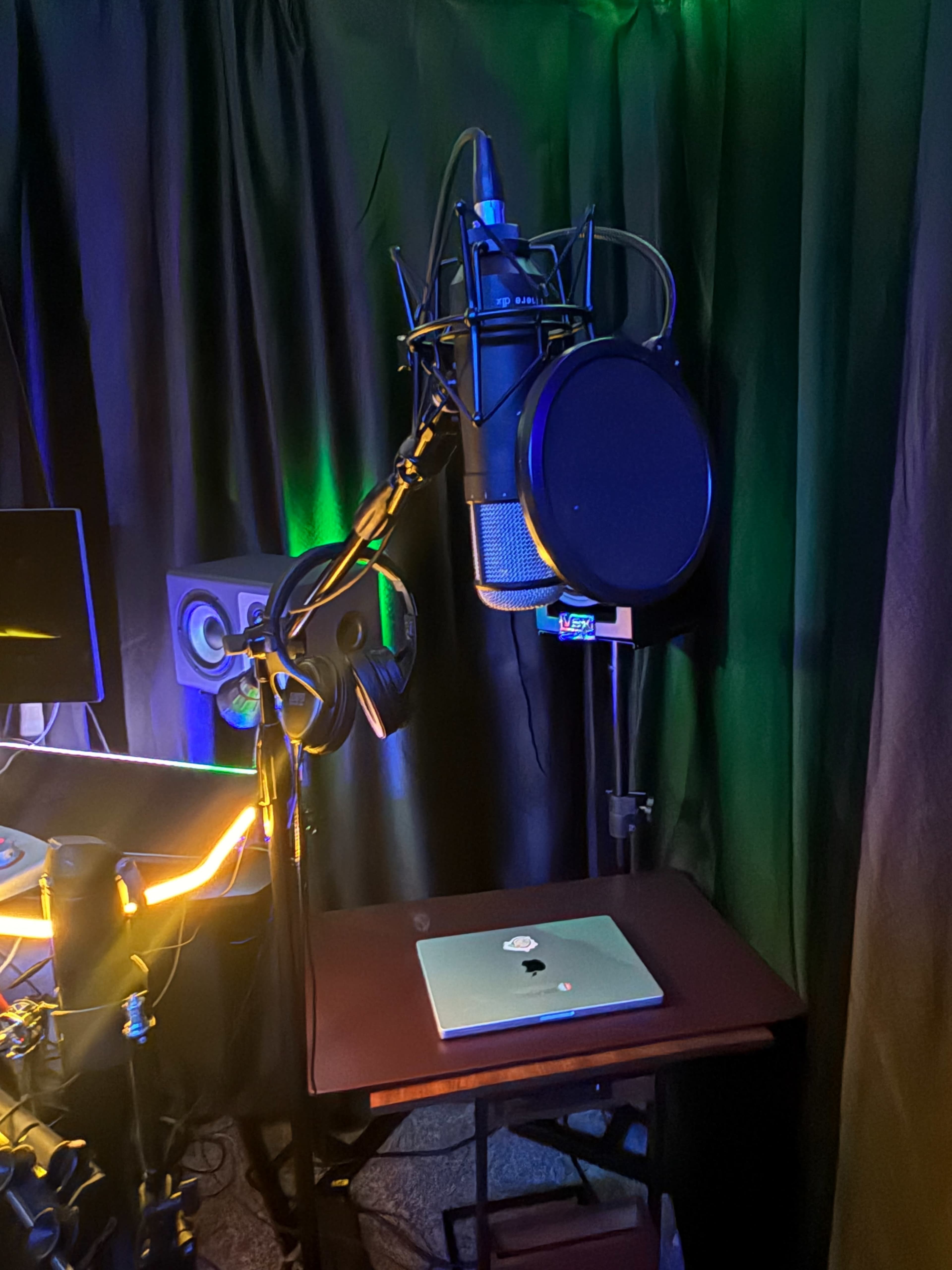The image shows a recording setup featuring a microphone on a stand, a laptop on a small table, and sound equipment in a dimly lit room with colorful lighting.