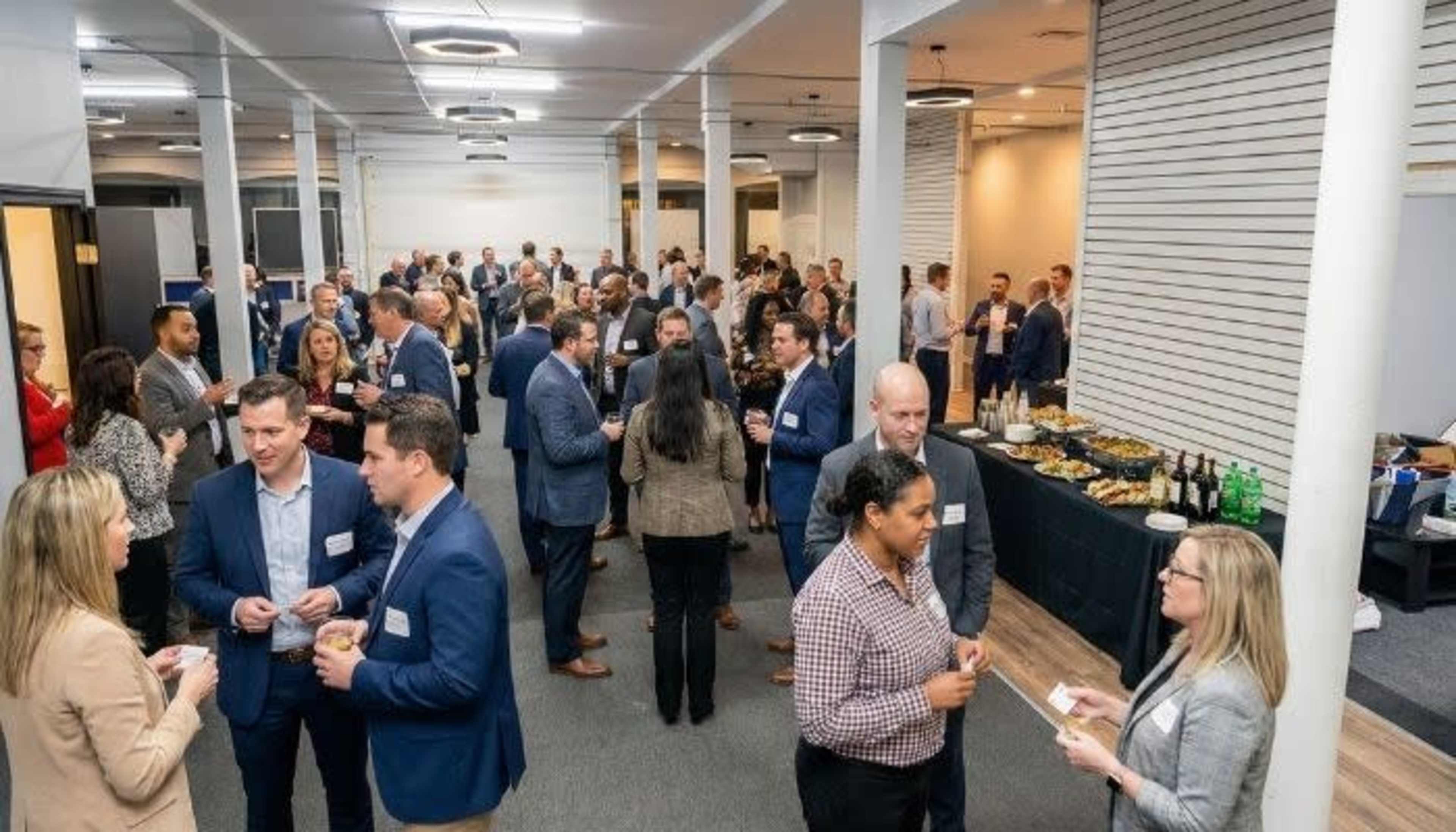 A crowded networking event is taking place in a well-lit room with attendees engaged in conversations and refreshments on display at a buffet table.