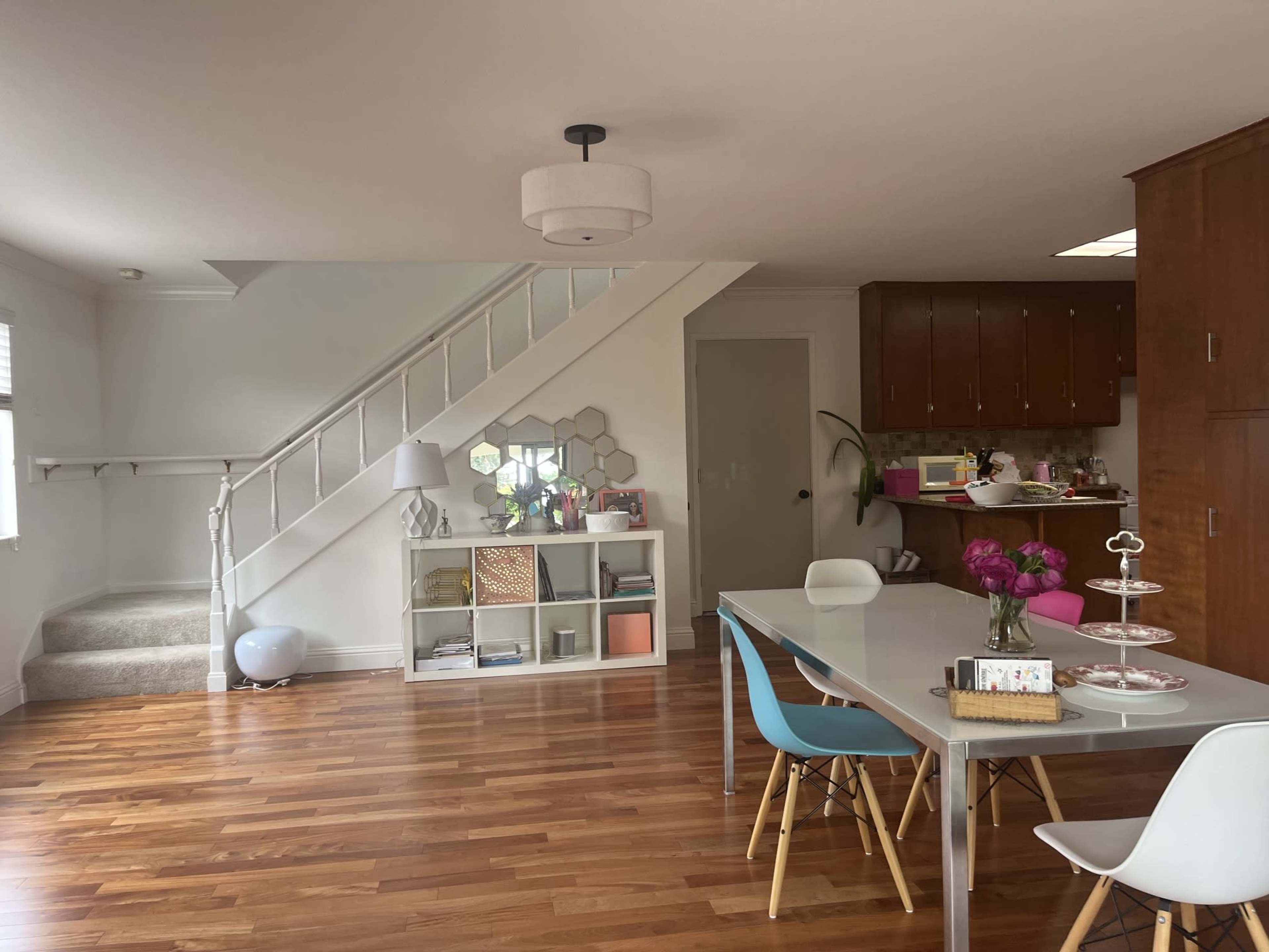 The image shows a modern living space featuring a dining area with a glass table, colorful chairs, and a staircase in the background, alongside a kitchen visible in the distance.