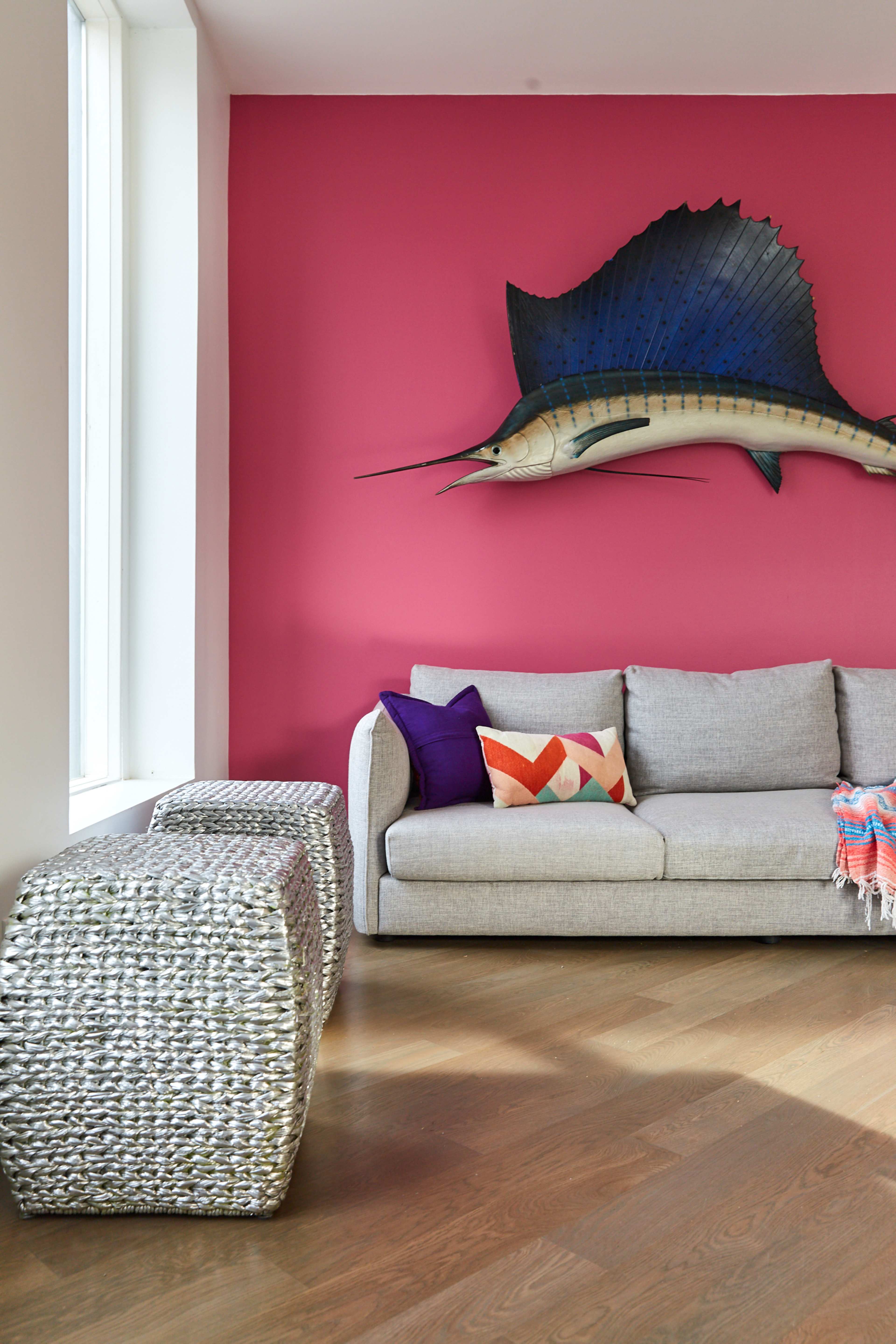 A gray sofa with decorative pillows is positioned against a bright pink wall, which features a large fish sculpture above it, alongside two woven side tables on the wooden floor.