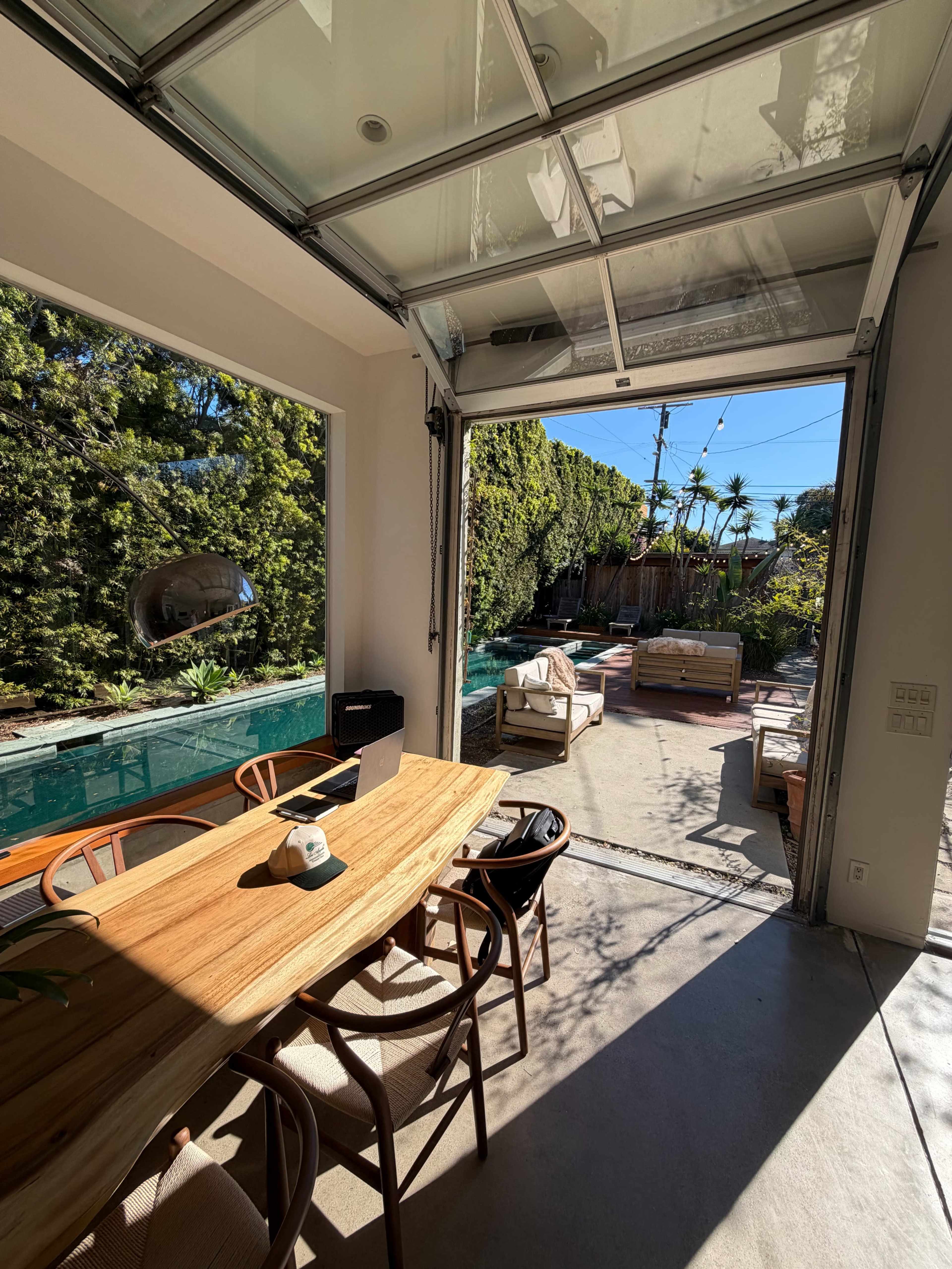 The image shows a bright indoor space with a large wooden table, overlooking a backyard area featuring a pool and lounge furniture through an open garage-style door.