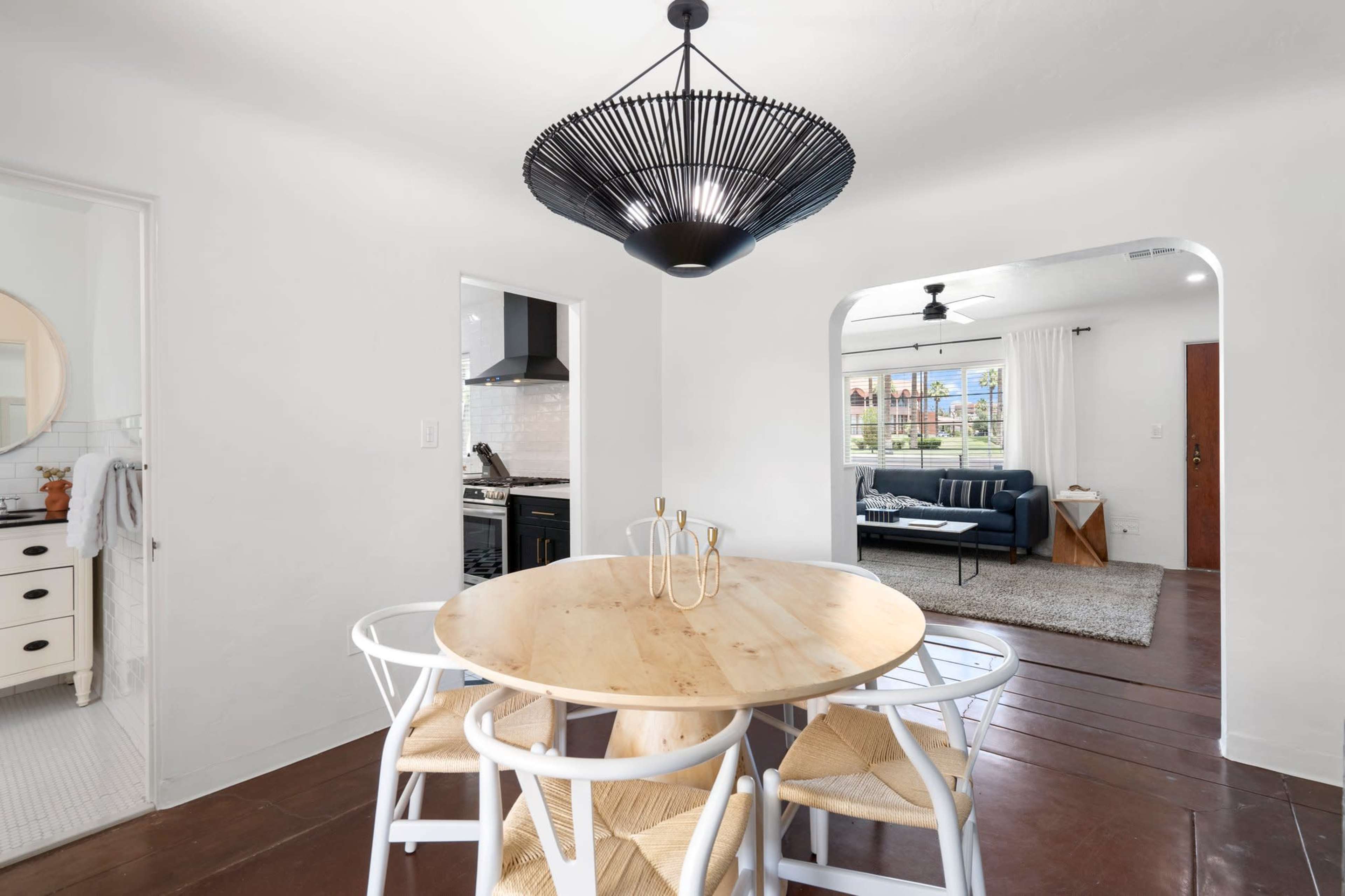 A round wooden dining table with white chairs is set in a brightly lit room featuring an open layout, leading to a modern kitchen and living area.