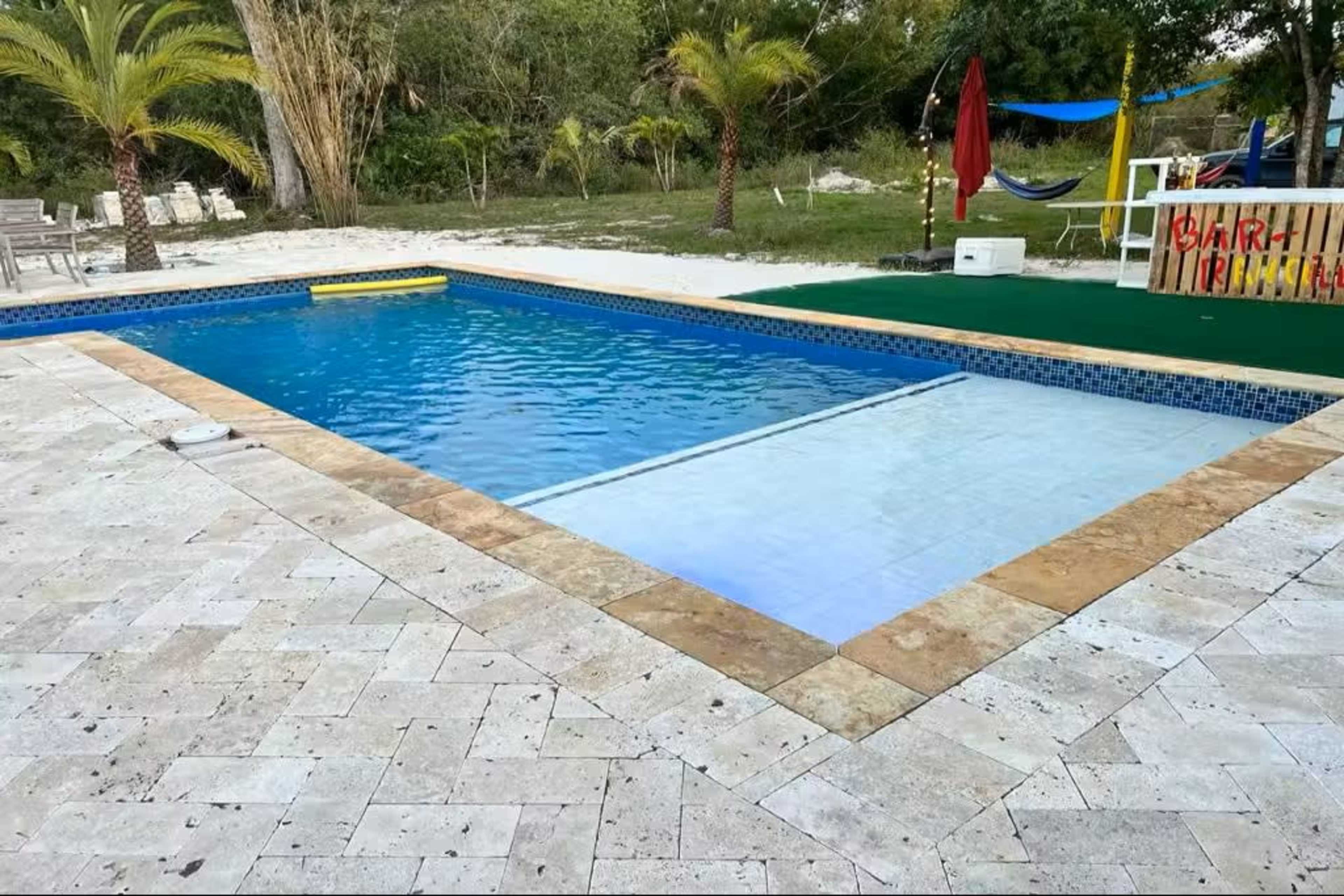 The image shows a rectangular swimming pool surrounded by tile and palm trees, with a grassy area and lounge chairs nearby.