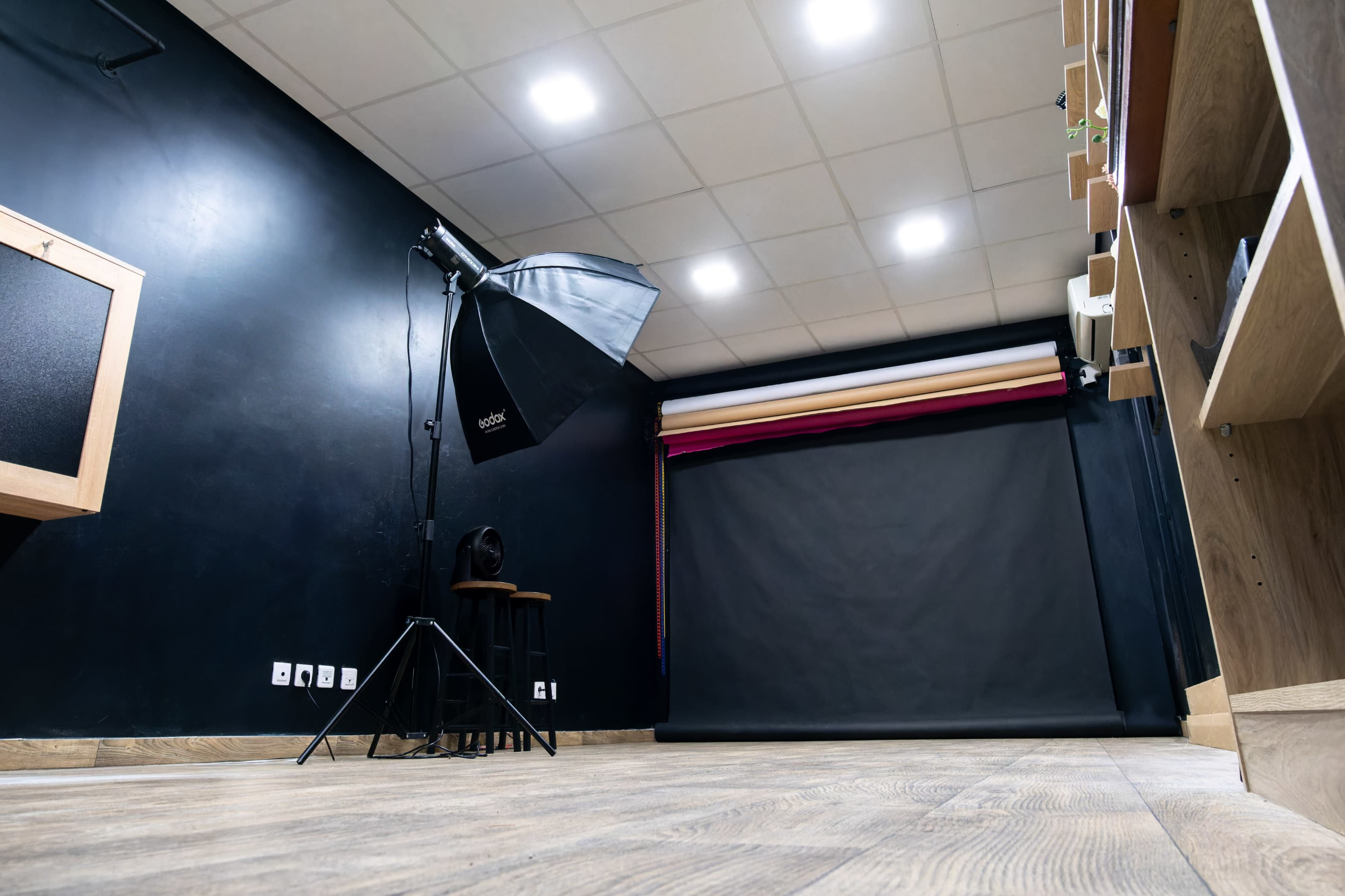 The image shows a photography studio with a black backdrop, lighting equipment, and wooden flooring.
