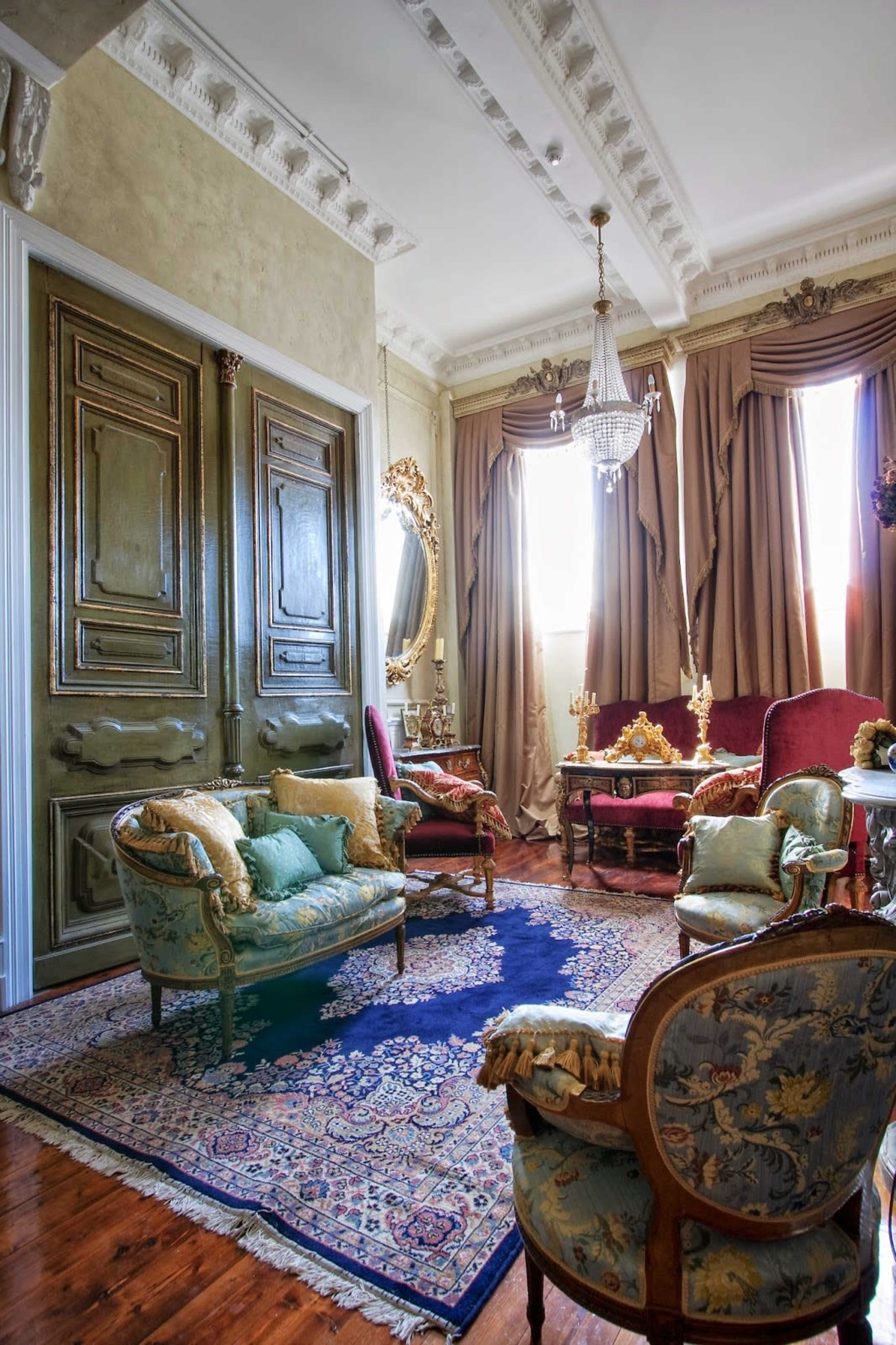 The room features ornate wooden doors, elegant seating arrangements, and large drapes with a chandelier illuminating the space.