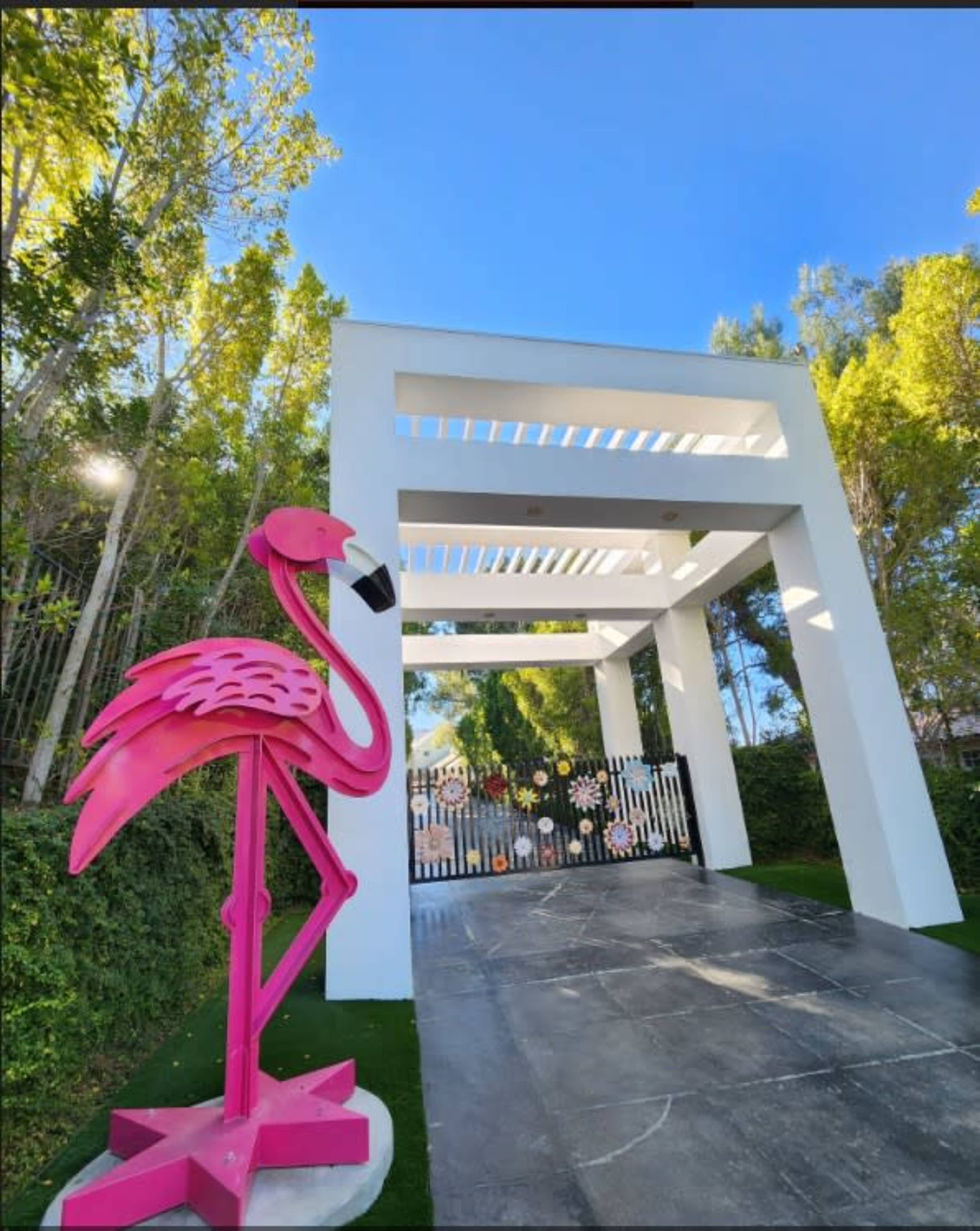 A large pink flamingo statue stands beside a modern entrance structure with a white pergola and a decorative gate.