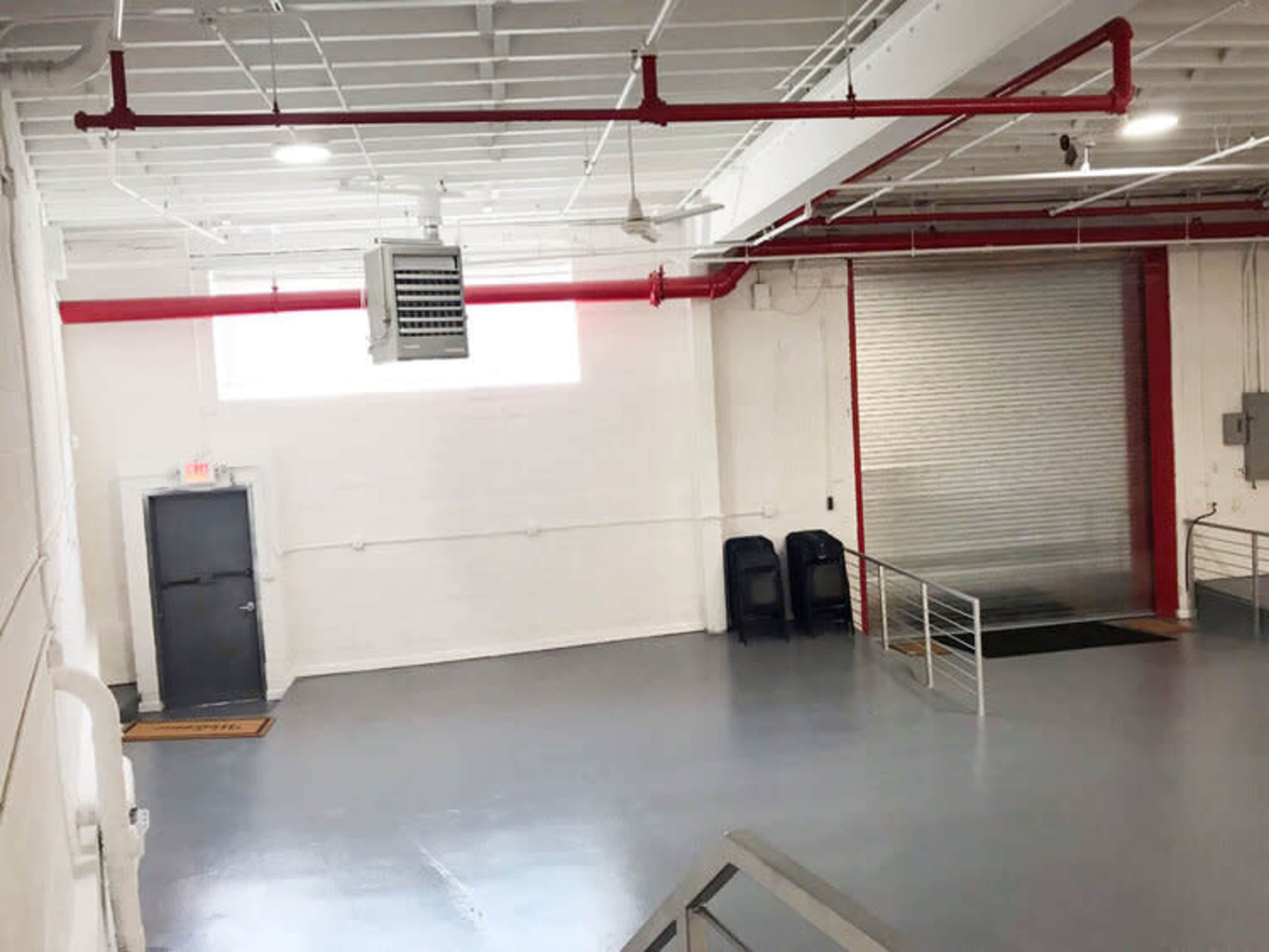 An empty industrial interior space with white walls, a large roll-up door, and two black chairs near a metal ramp.
